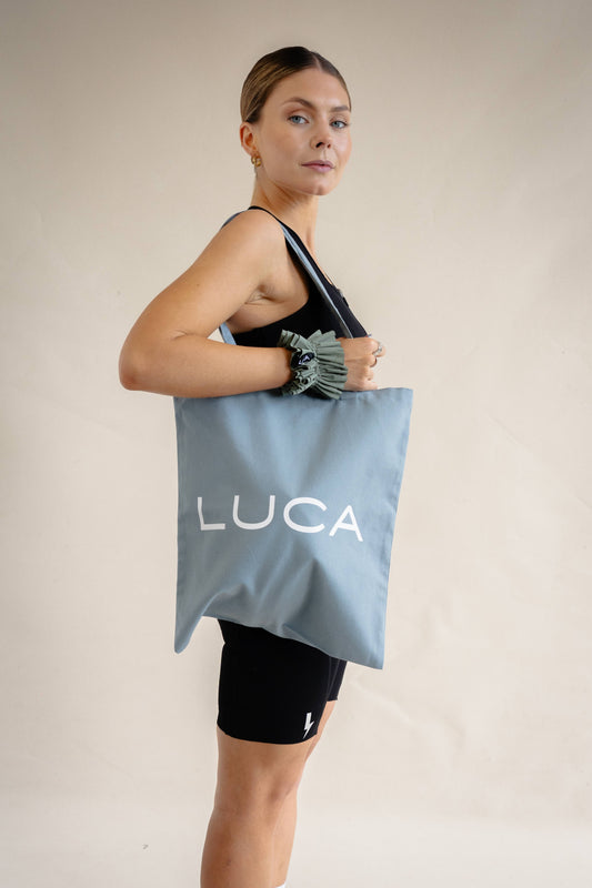 A side profile lifestyle shot of a woman against a cream background wearing a matcha green ruffled fabric scrunchie on her wrist while carrying a slate blue LUCA® tote bag.