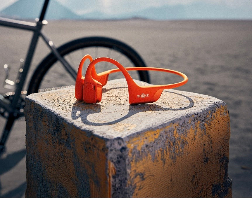 Close-up-of-bright-orange-Shokz- OpenRun-bone-conduction- headphones-resting-on-a-weathered-concrete-block-with-a- road-bike-and-mountain-landscape- in-the-out-of-focus-background