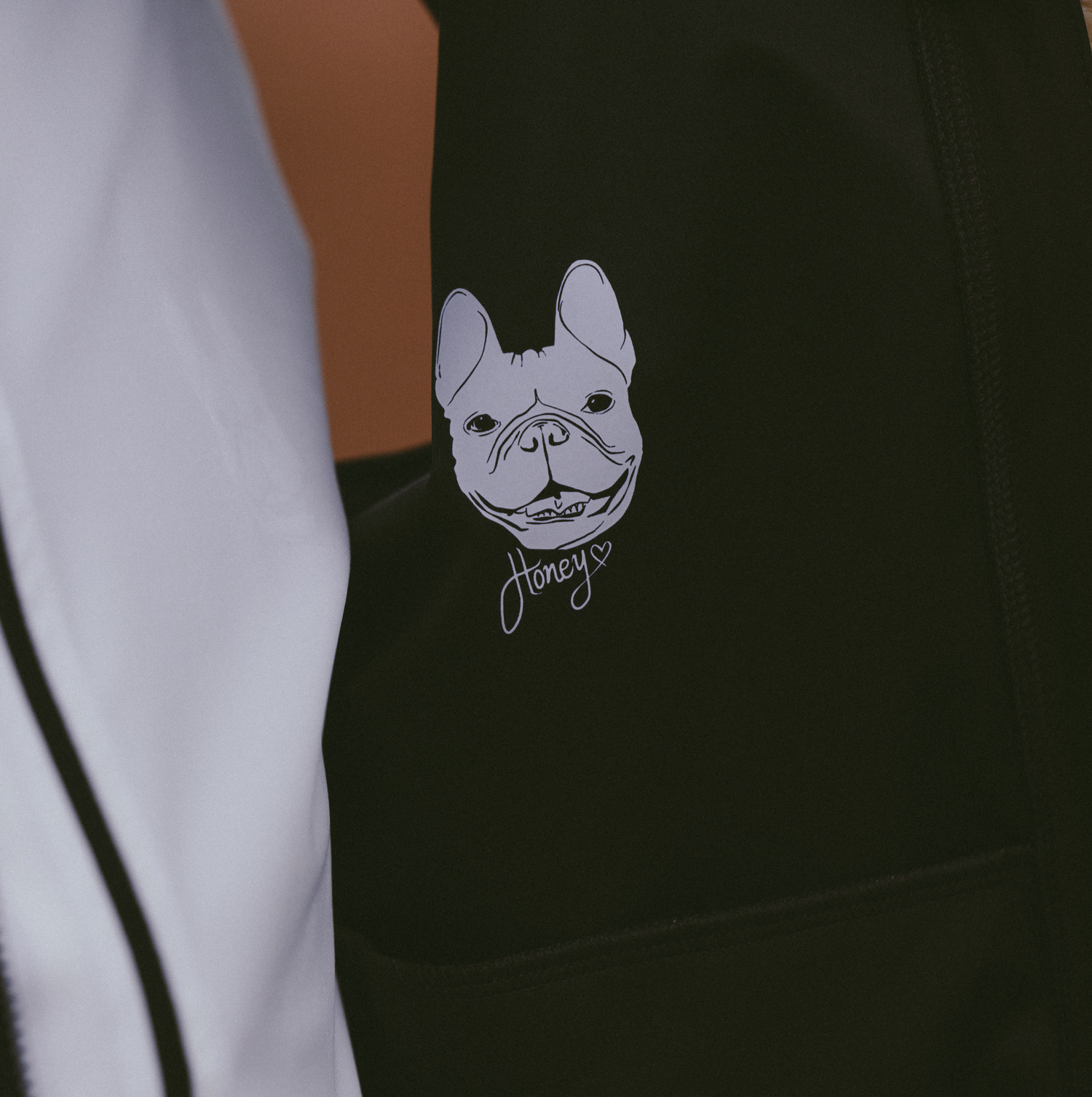 Close-up-of-a-black-garment-likely-a- Ascend-Gilet-featuring-a-white-graphic-print-of-a-happy-French- Bulldog's-face-Below-the-dog's-face-the-word-Honey-is-written-in-a- delicate-white-script-with-a-small-heart-outline-A-section-of-a-white- garment-possibly-with-black-stripes-is-visible-on-the-left-side