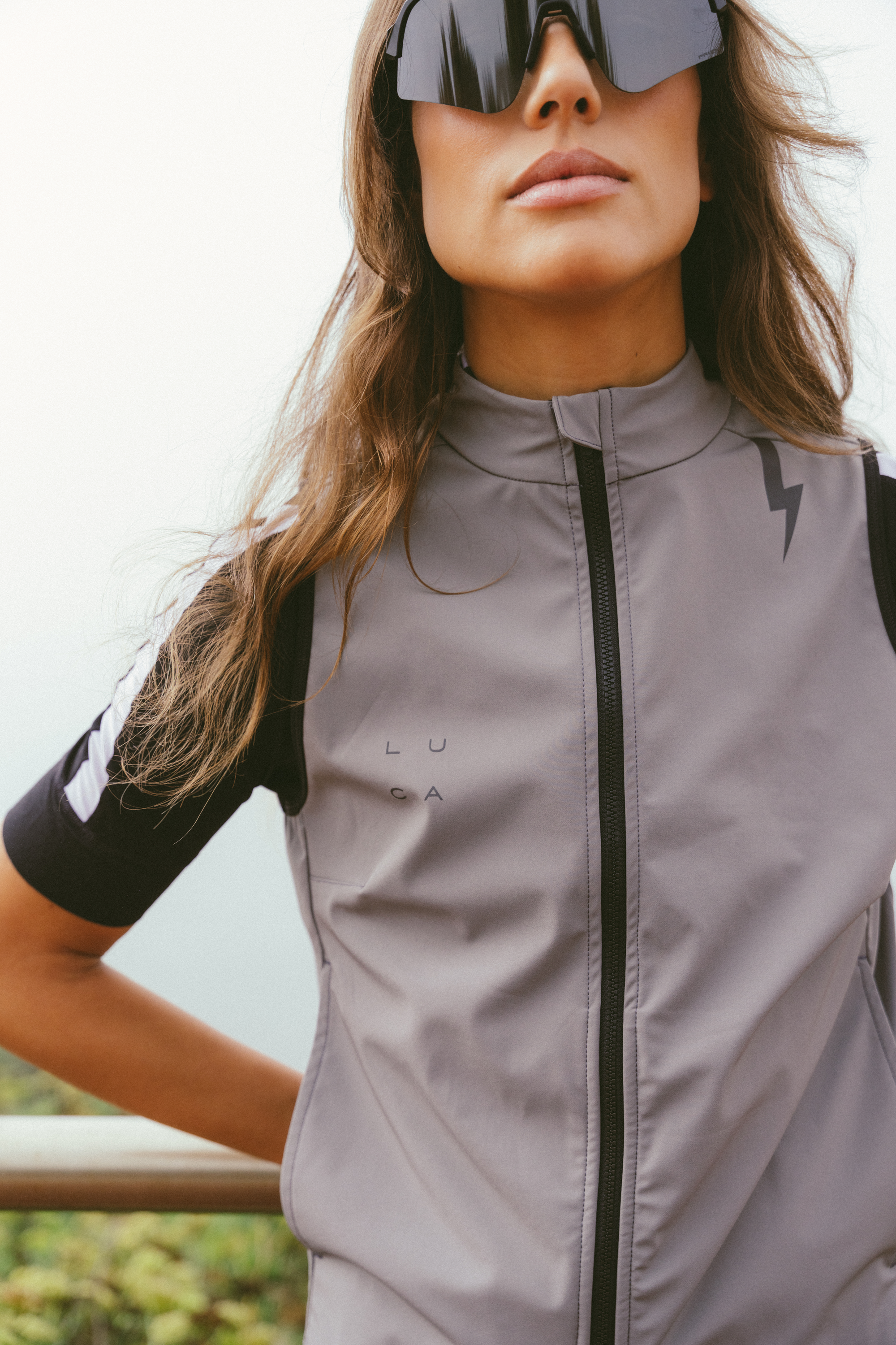 Close-up-of-a-woman-wearing-a- light-gray-athletic-Ascend-Gilet-with-a-black-full-zip-closure-and-a- high-collar-The-vest-features-a- small-subtle-black-LUCA-logo-on-the-chest-and-a-contrasting-black- lightning-bolt-graphic-on-the-right- shoulder-She-is-wearing-black- short-sleeved-cycling-wear- underneath-which-has-white-stripes-on-the-sleeve-and-is- accessorized-with-black-wrap-around-cycling-sunglasses