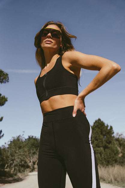 A-woman-posing-outdoors-viewed- from-a-low-angle-against-a-clear- blue-sky-She-is-wearing-a-black- zipped-cycling-bra-and-matching- high-waisted-leggings-with-a-thick- white-vertical-stripe-on-the-side- She-is-also-wearing-reflective-shield-sunglasses-with-a-yellow- frame-Focus-on-women's-black- activewear-set-running-outfit-and- high-waist-leggings