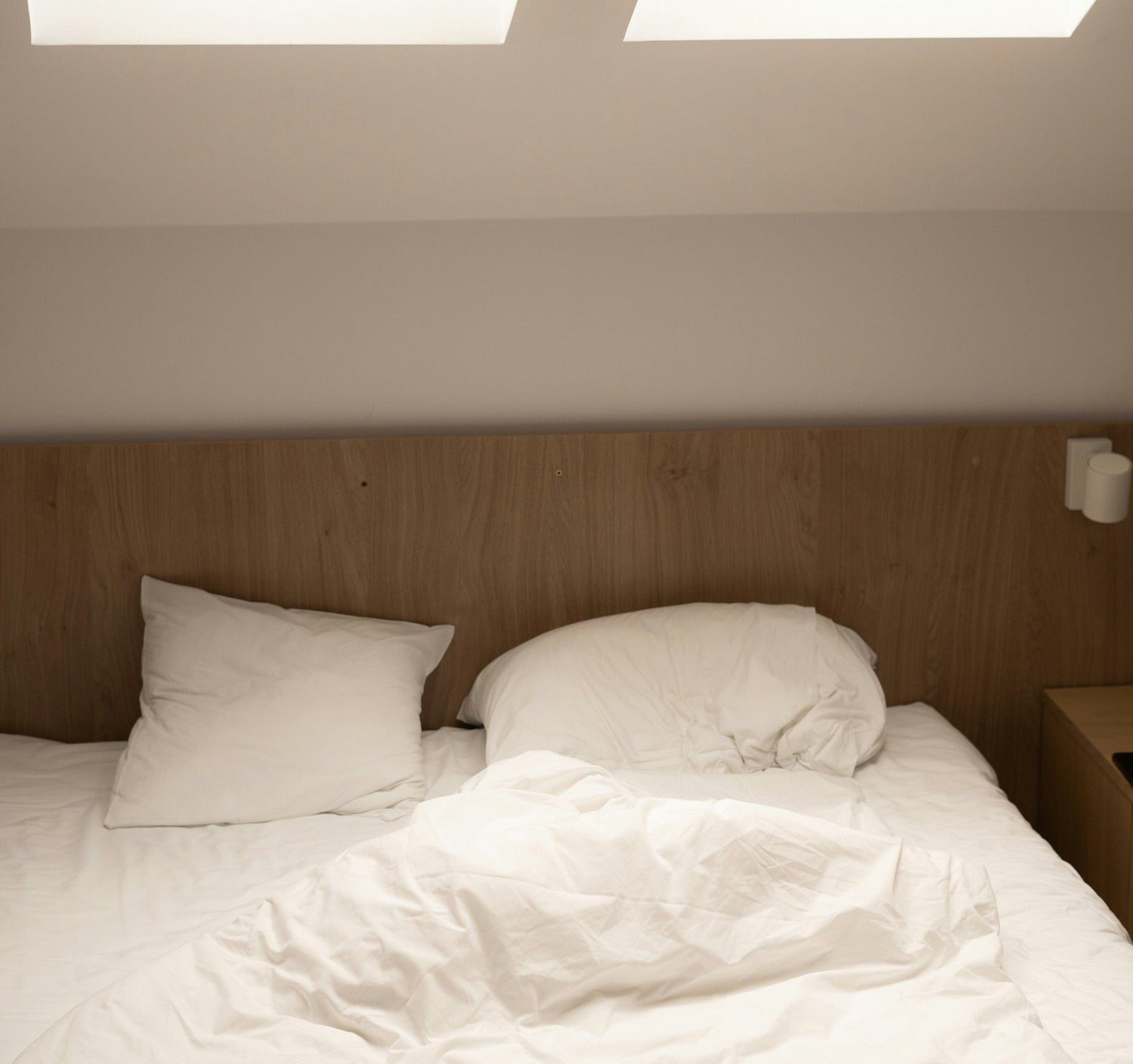 Close-up-of-an-unmade-bed-with-a- messy-white-duvet-two-fluffy-white- pillows-and-a-light-wood-headboard-suggesting-comfort