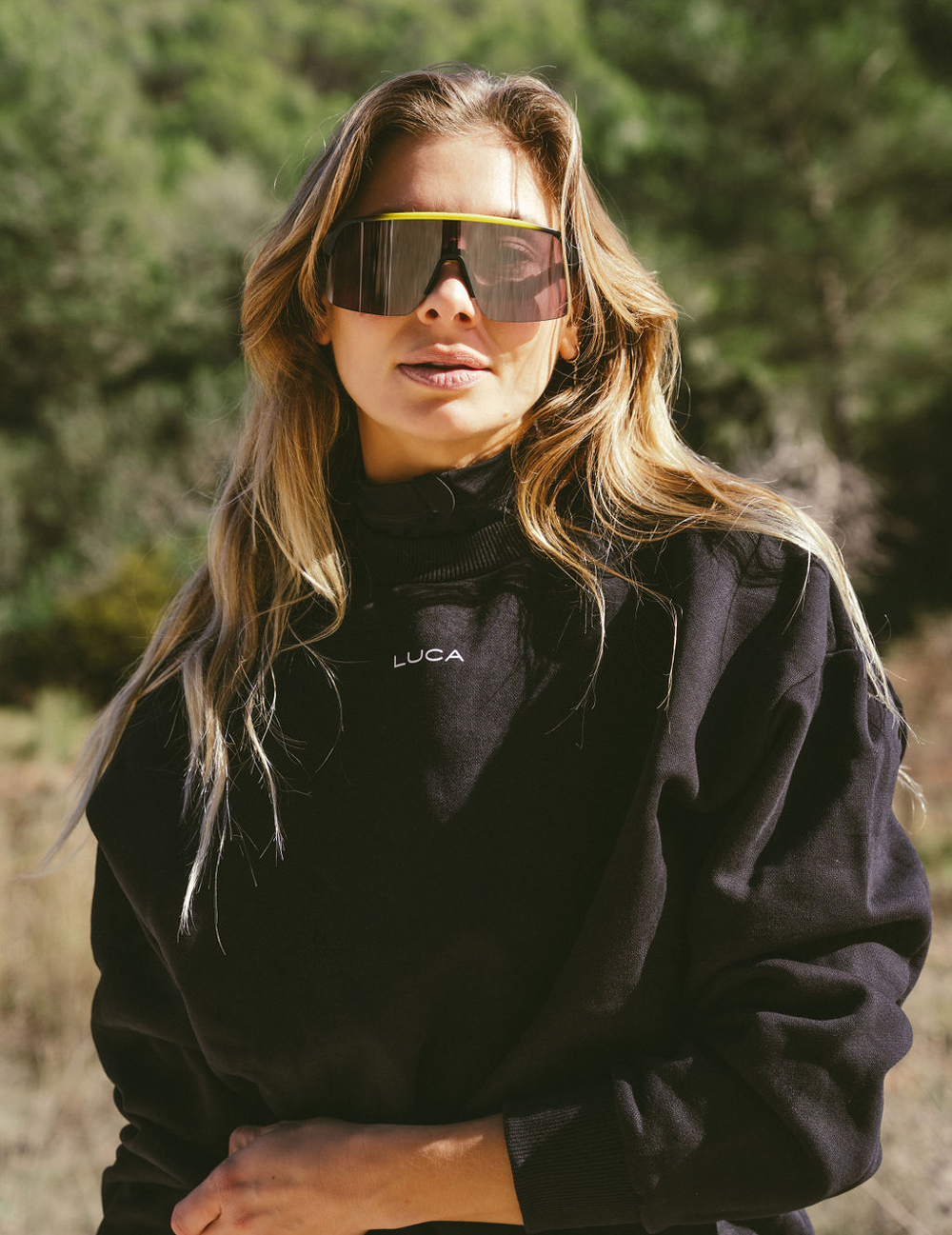 A-close-up-outdoor-photograph-of-a-woman-with-long-wavy-blonde- hair-wearing-a-black-high-neck- shirt-The-brand-name-"LUCA"-is- printed-in-white-text-on-the-chest- She-is-also-wearing-oversized- performance-shield-sunglasses-with-mirrored-lenses-and-a- distinctive-neon-yellow-accent-along-the-top-frame-The- background-is-blurred-showing-a- sunny-wooded-brush-filled- landscape