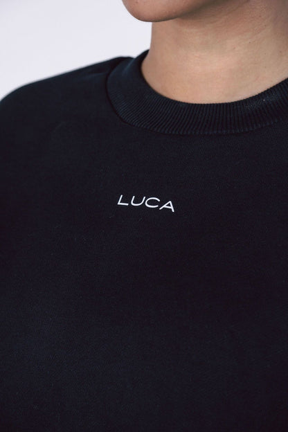 A close-up-detail-shot-of-a-black- crewneck-sweatshirt-showing-the- white-embroidered-LUCA-brand-logo- centered-below-the-neckline-Focus-on-black-crewneck-sweater- embroidered-logo-apparel-unisex- casual-wear-and-simple-modern- sweatshirt