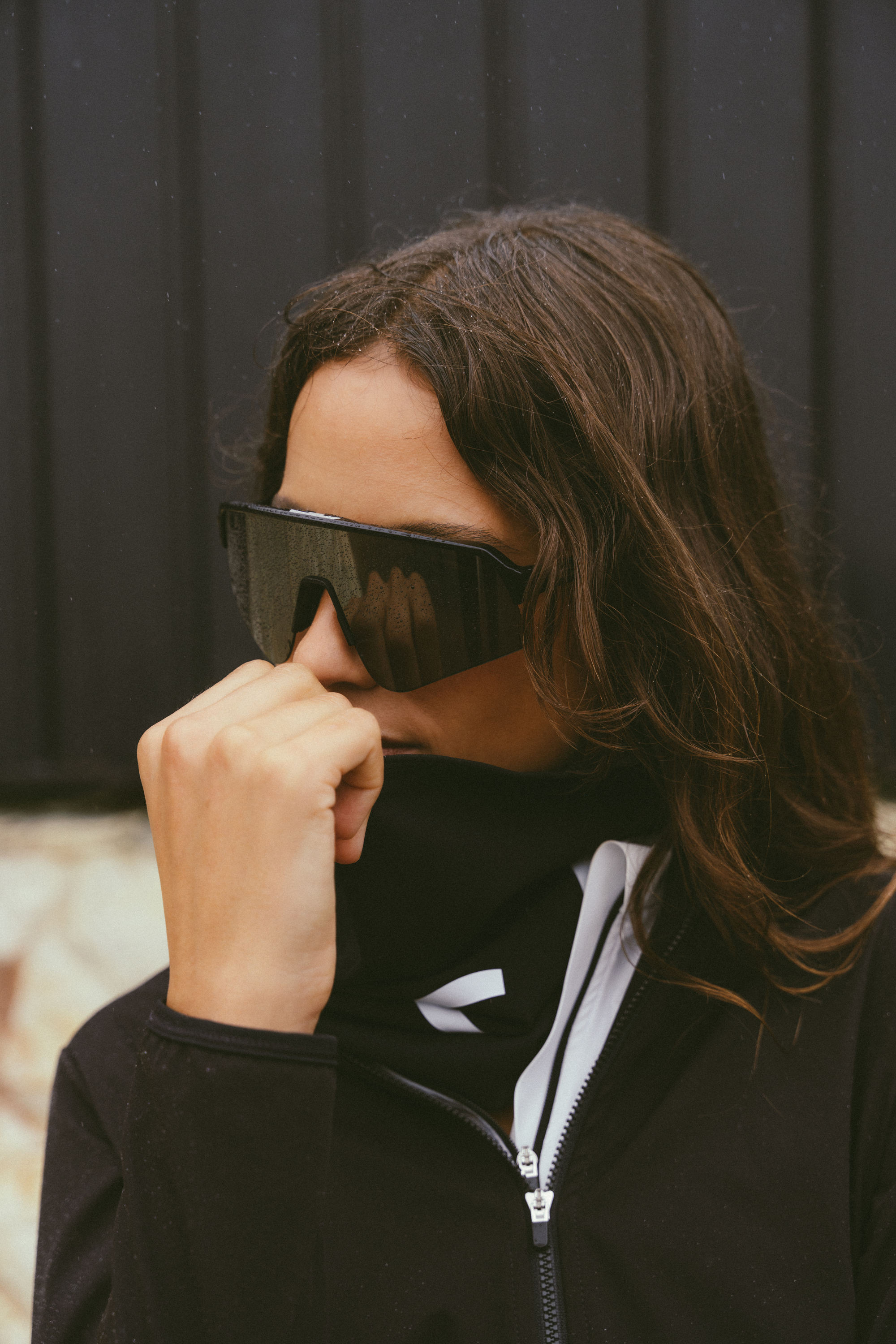 Close-up-of-a-person-wearing-large-black-futuristic-wrap-around- cycling-shield-sunglasses-They-are- wearing-a-black-high-collar- performance-jacket-with-Flow-Buff- with-a-white-visible-zipper-tape-and-a-white-stylized-logo- resembling-centered-on-the-chest- below-the-collar-The-person-is- covering-their-mouth-with-their-hand-while-posing-outdoors