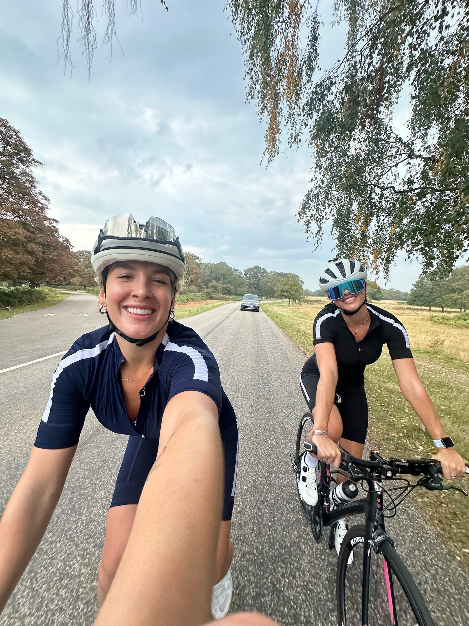 Two-happy-female-cyclists-in- matching-navy-and-black-short-sleeve-cycling-kits-taking-a-selfie- while-riding-their-road-bikes