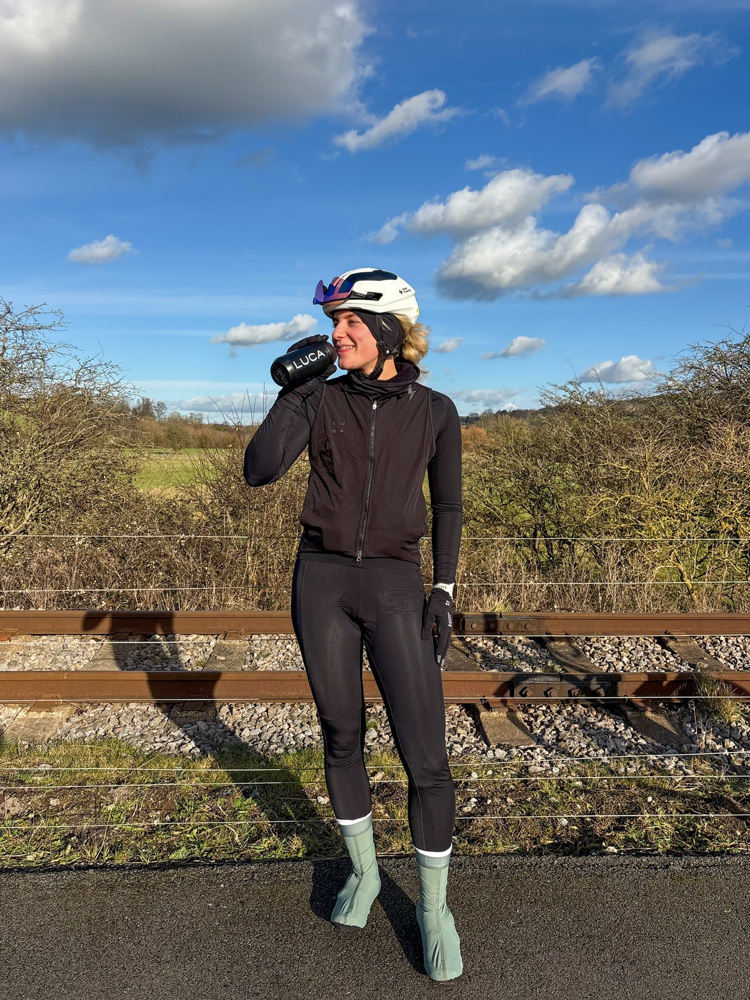 Female-cyclist-drinking-from-a-black- water-bottle-while-standing-outdoors-in-black-leggings-a-vest- and-a-white-helmet-on-a-sunny-day