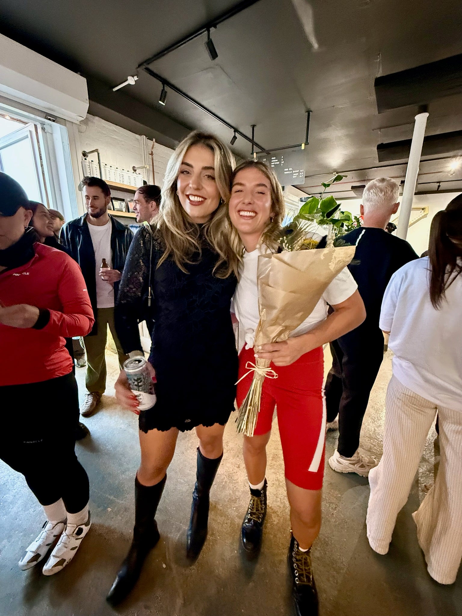 A-candid-indoor-photograph-of-two- young-women-smiling-and-posing- together-in-a-crowded-setting-likely-a- shop-event-party-The-woman-on-the- left-is-dressed-in-a-black-lace-dress- and-high-black-boots-The-woman-on- the-right-is-holding-a-bouquet-of- flowers-and-wearing-red-cycling- shorts-paired-with-a- simple-white-t-shirt-black-lace-up- boots-and-white-socks-The-image- captures-a-contrast-of-styles-and- emphasizes-the-brand-community- and-celebratory-atmosphere