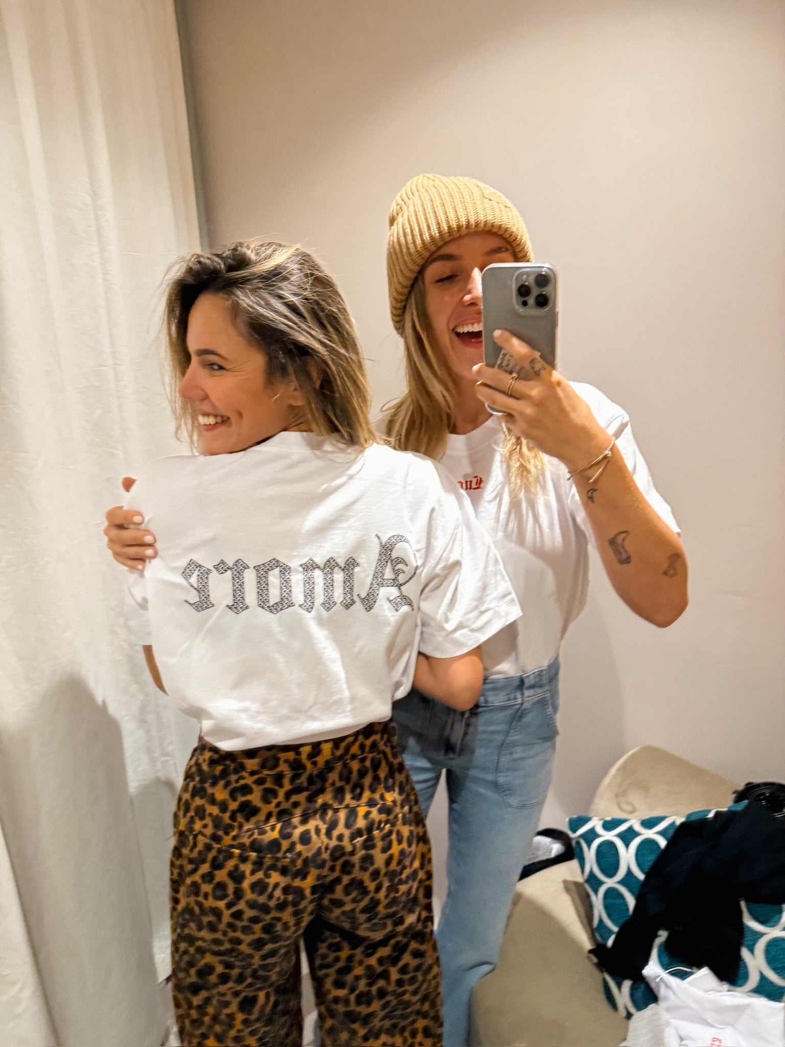 Two-smiling-women-taking-a-selfie- in-a-fitting-room-The-woman-facing-away-wears-a-white-t-shirt- with-a-rhinestone-studded-text- design-on-the-back-text-is-reversed-but-appears-to-read- 'AMARÉ-paired-with-leopard-print- pants-The-other-woman-wears-a-tan-beanie-and-light-wash-jeans