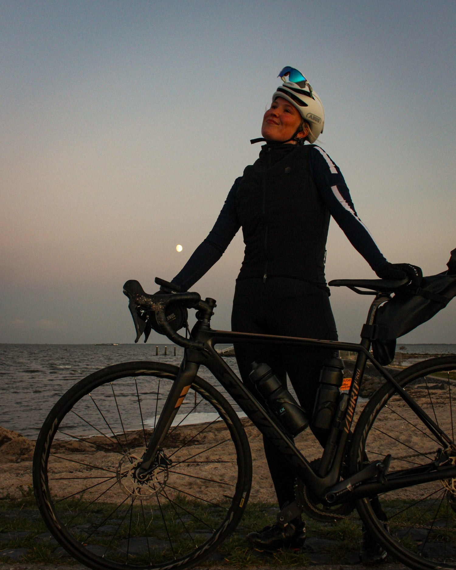 Female-cyclist-standing-with-her- black-road-bike-at-sunset-by-the- ocean-wearing-a-helmet-and-black- cycling-clothes