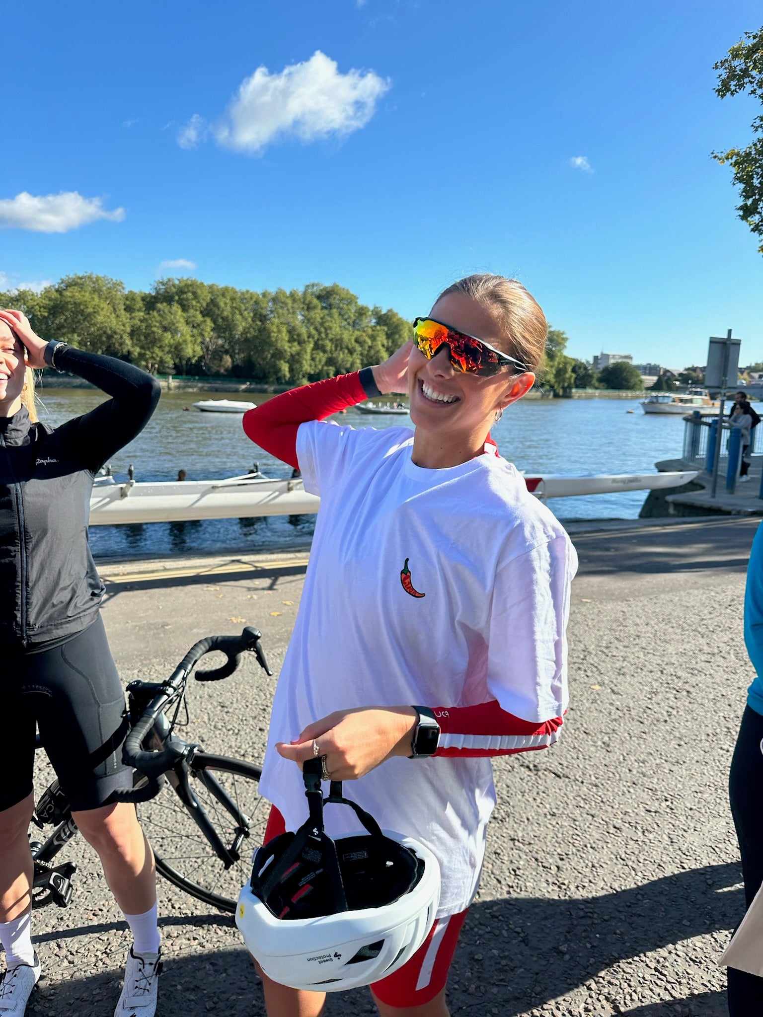 Cyclist-smiling-outdoors-next-to-a-river-on-a-sunny-day-wearing-a-white- t-shirt-with-a-chili-graphic-reflective-sunglasses-and-holding-a- helmet