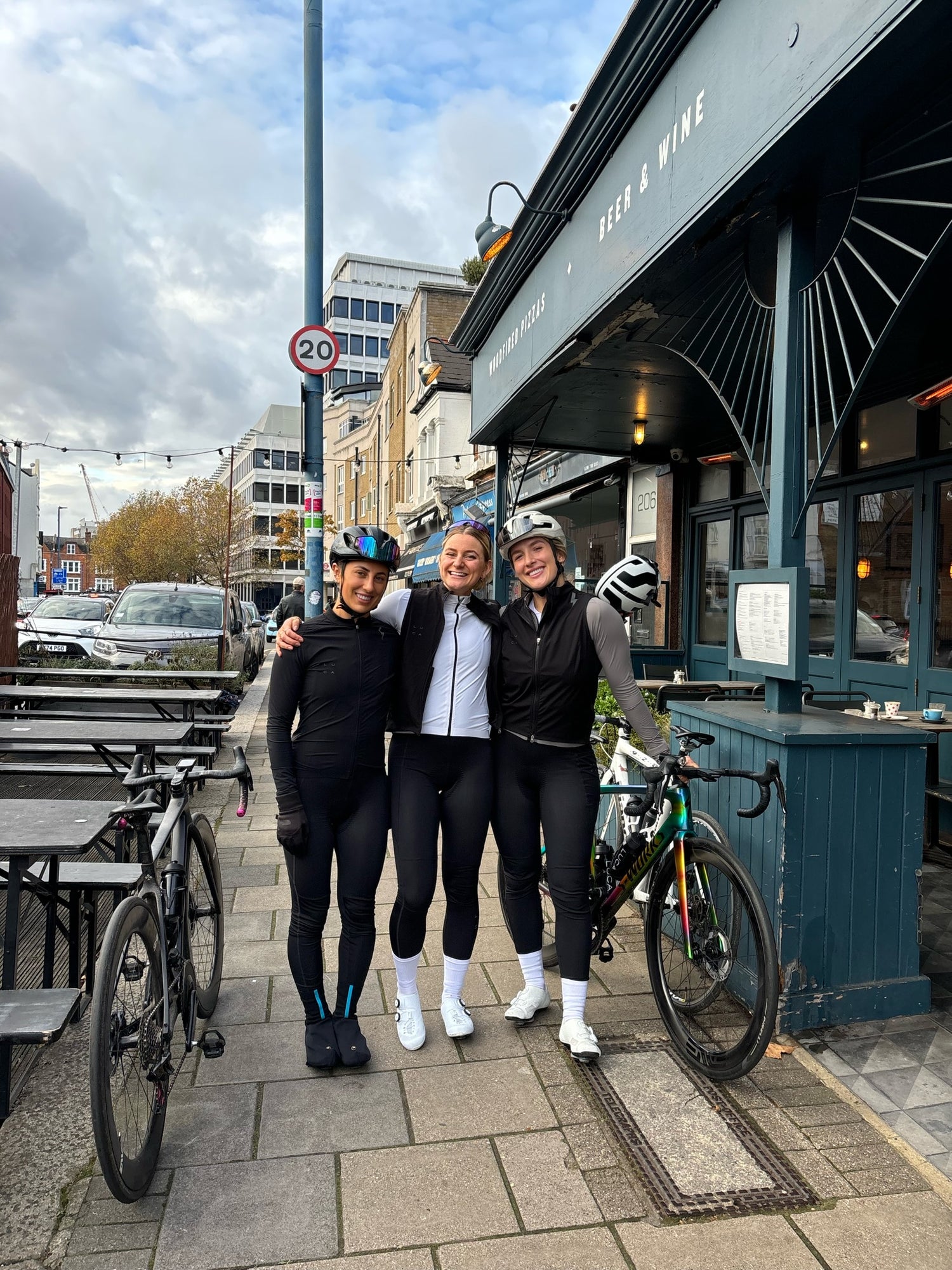 Three-female-cyclists-smiling-in- cycling-gear-outside-a-Beer-&-Wine-pub-in-an-urban-street-setting