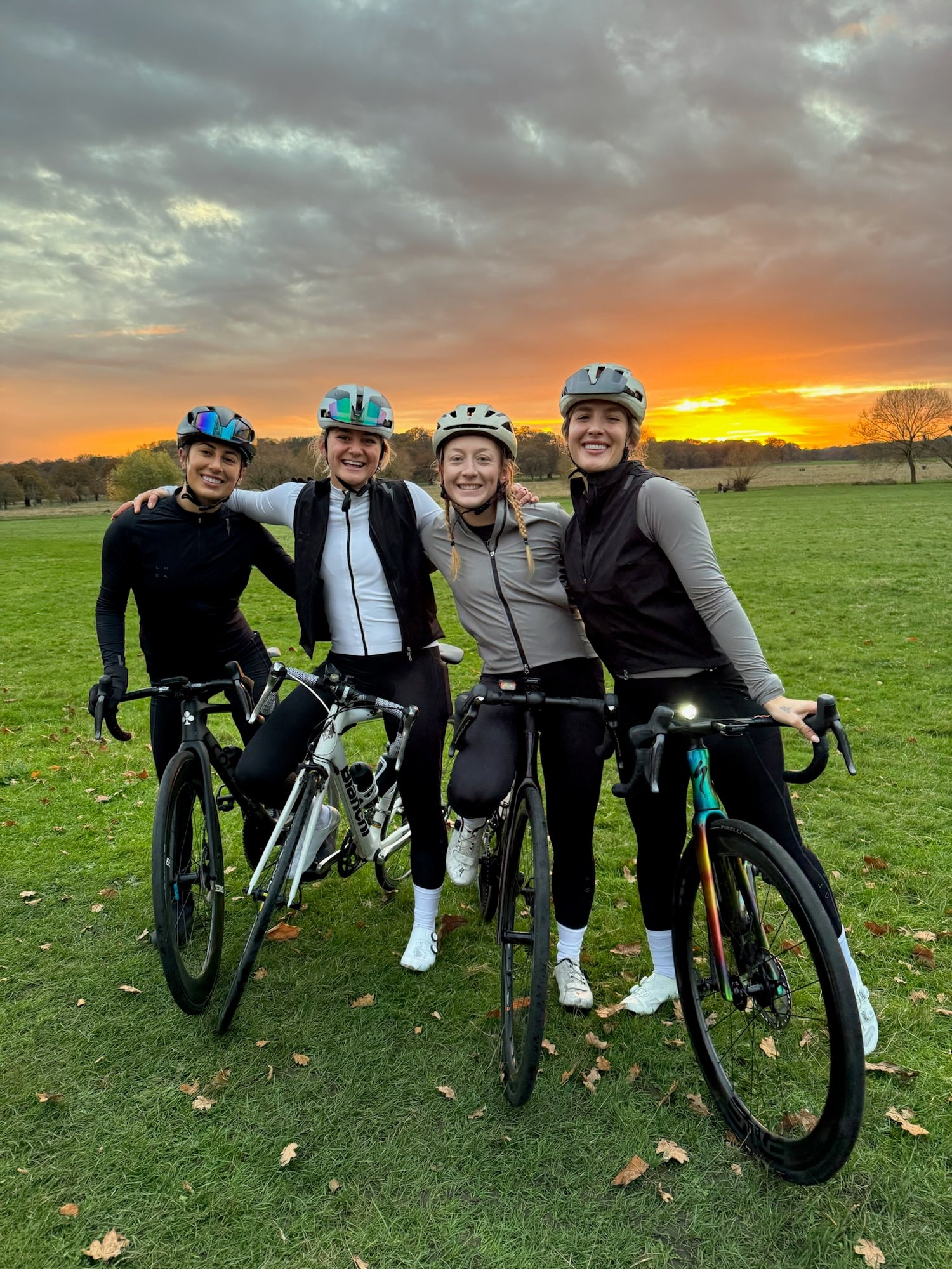 Group-photo-of-four-women-cyclists-in-black-and-white-gear-and-helmets-standing-with-their- bikes-in-a-park-at-sunset