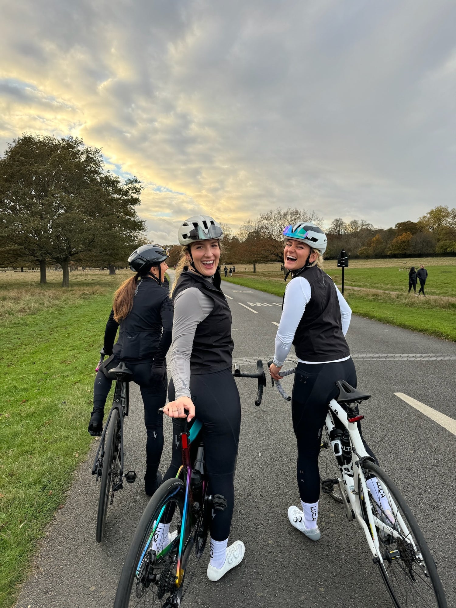Three-happy-female-cyclists-in- black-vests-and-helmets-pausing- their-road-bikes-on-an-asphalt-path- in-a-large-nature-area