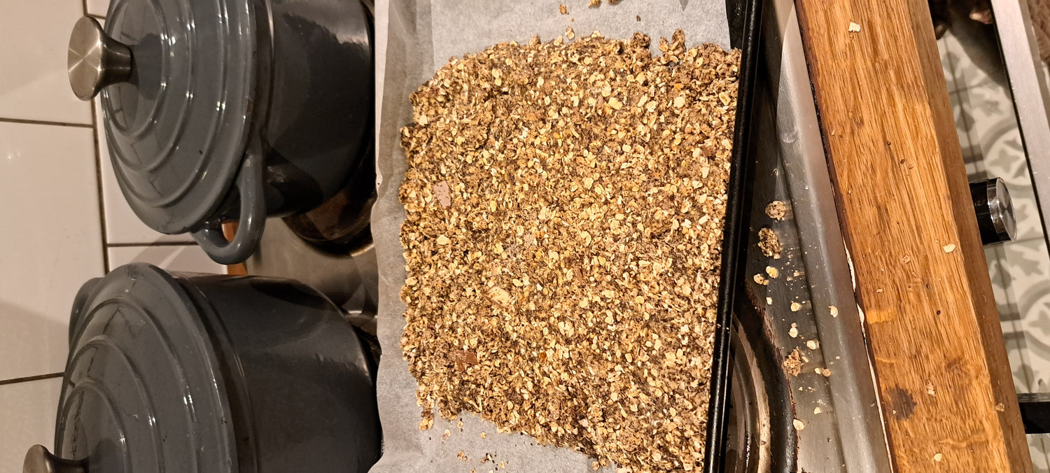 Close-up-of-golden-brown-textured- granola-flapjack-mixture-spread-out- on-parchment-paper-on-a-baking- sheet-ready-for-the-oven-Two-grey- Dutch-oven-pots-are-visible-in-the- background