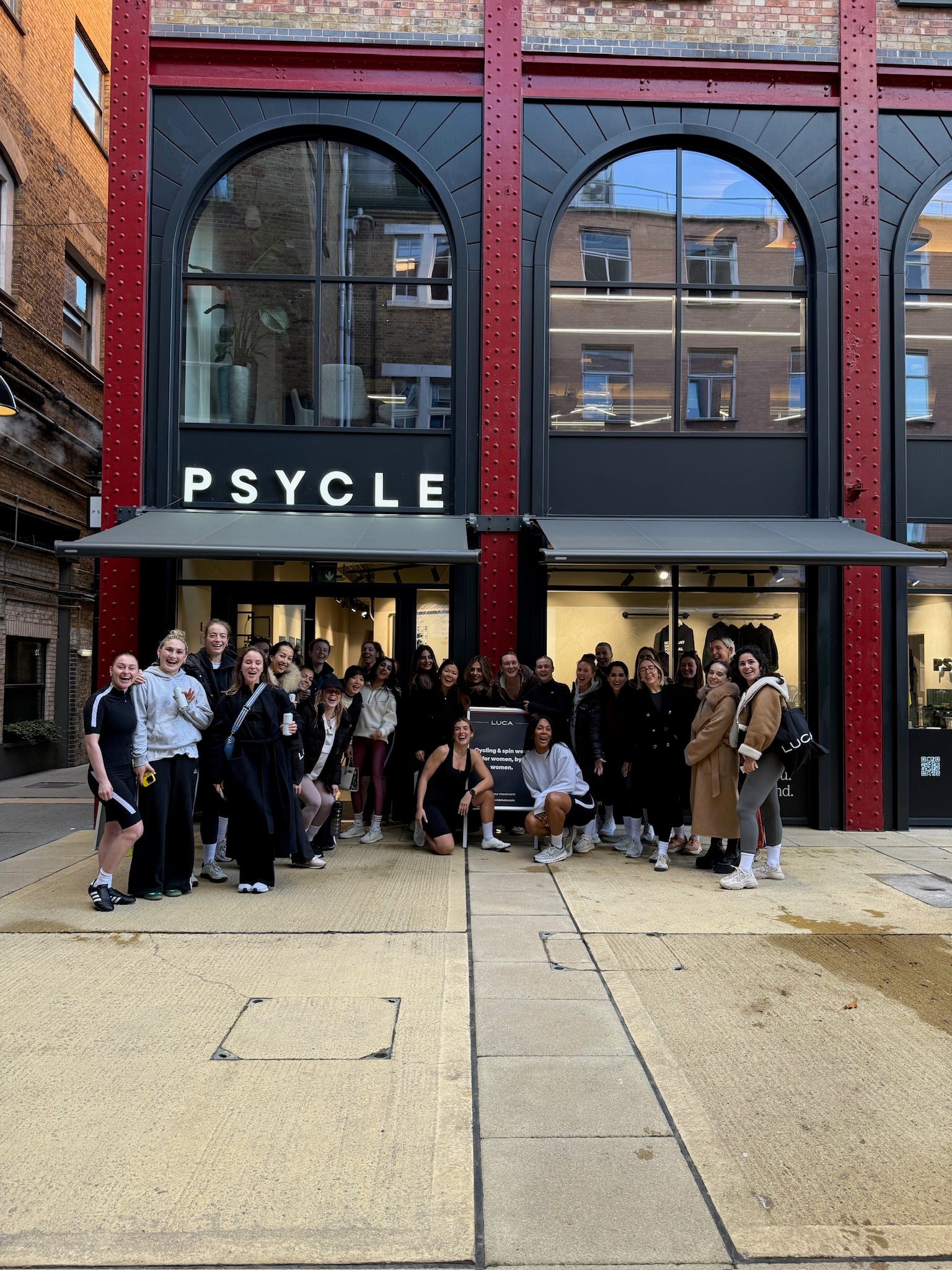 Big-group-of-people-standing-in- front-of-a-brick-building-with-a-sign-that-says-'PSYLE,'-likely-after-a-fitness-class