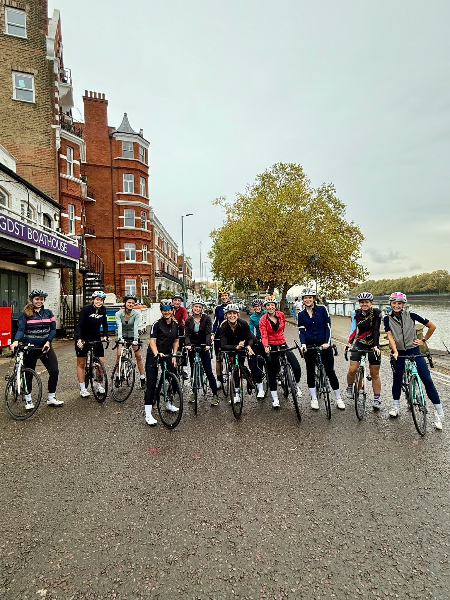 Group-of-people-on-road-bikes- standing-together-on-a-waterfront- path-in-front-of-a-tall-red-brick- building