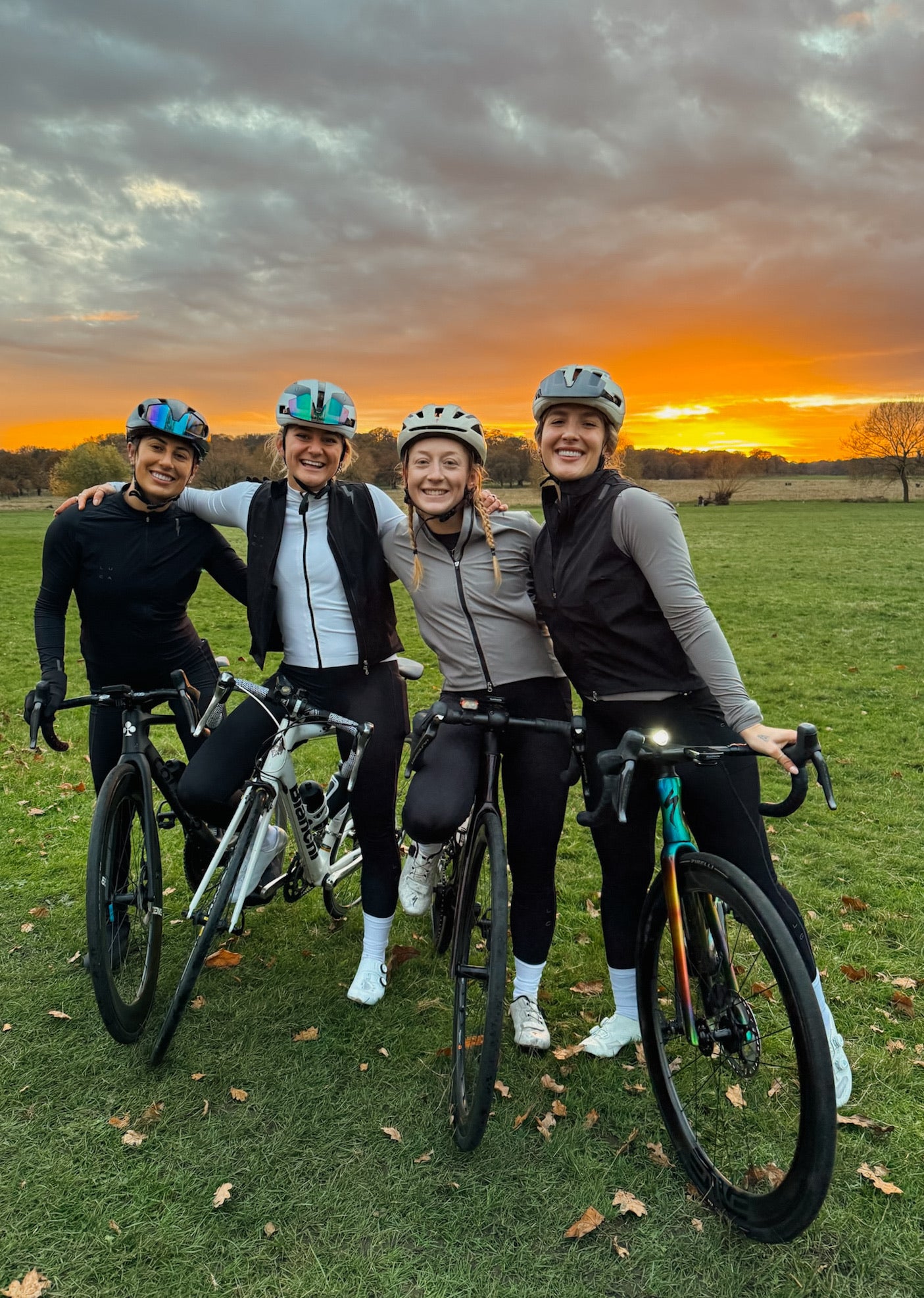 Four-women-cyclists-posing-with-their-bikes-in-a-grassy-park-at-sunset-all-wearing-helmets-and- athletic-tops