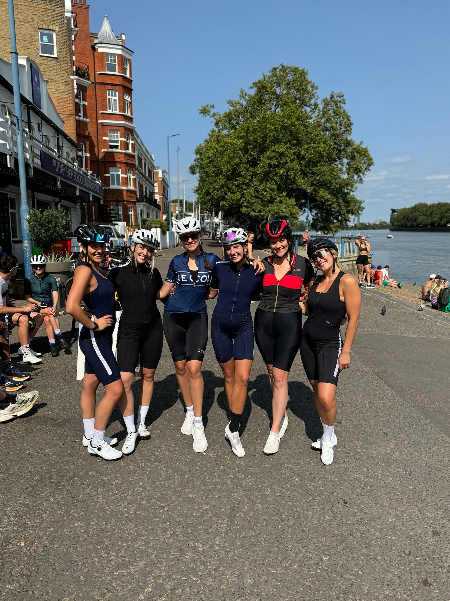 Group-of-six-happy-women-cyclists- posing-in-navy-blue-black-and-red- athletic-cycling-kits-next-to-a-river- on-a-sunny-day-with-classic-red- brick-architecture-in-the- background