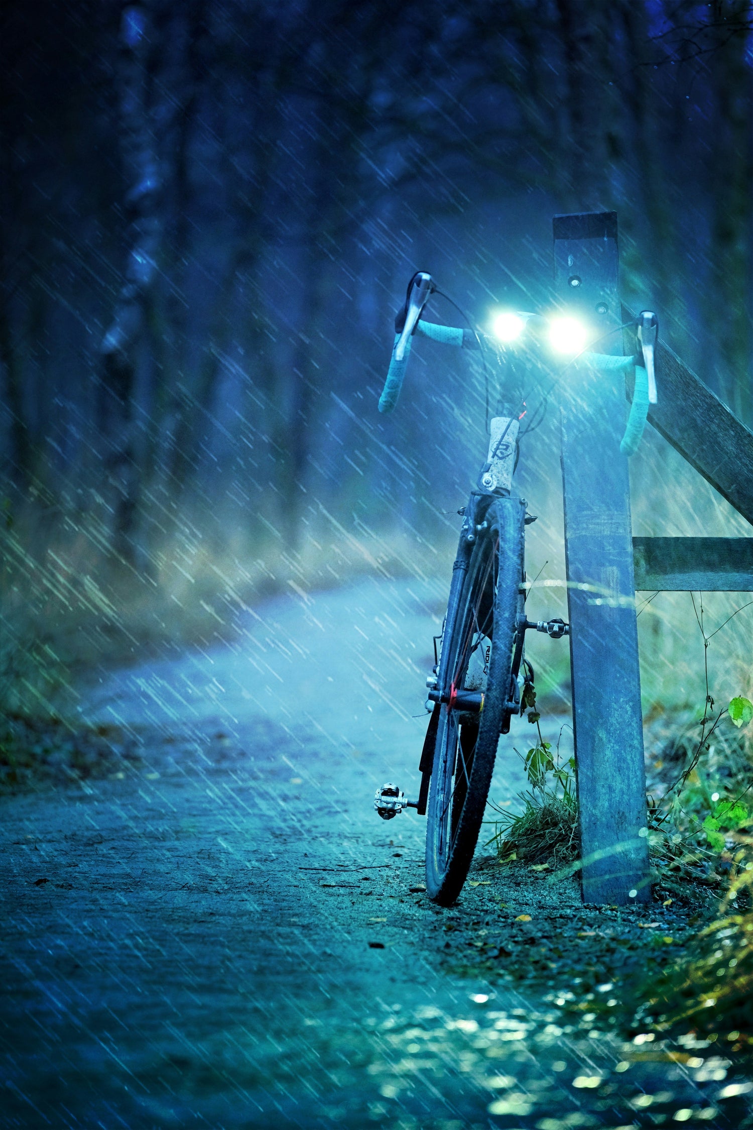 Bicycle-with-a-bright-front-light-parked-against-a-wooden-fence-post- on-a-wet-rainy-trail-at-night- dusk