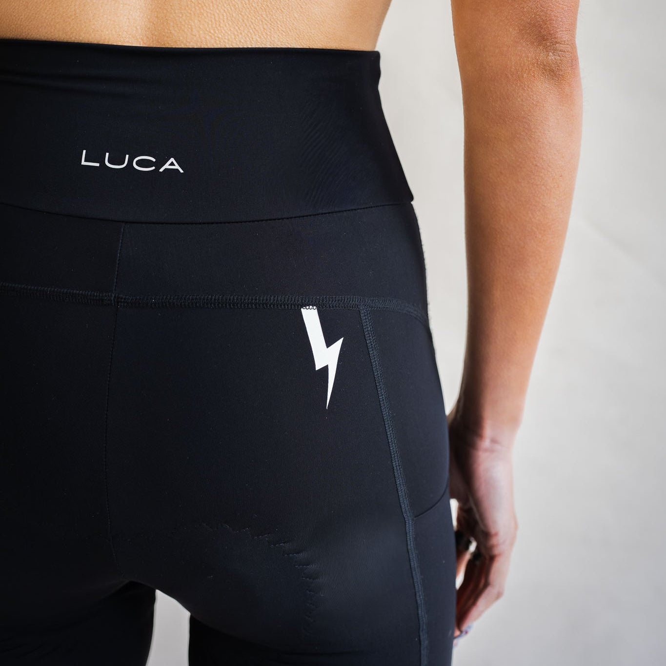 A close-up lifestyle shot from behind showing a model wearing high-waisted black LUCA cycling shorts. The image highlights branding placement, featuring a white LUCA® logo printed centered on the rear waistband and a distinctive white lightning bolt graphic on the upper right hip area