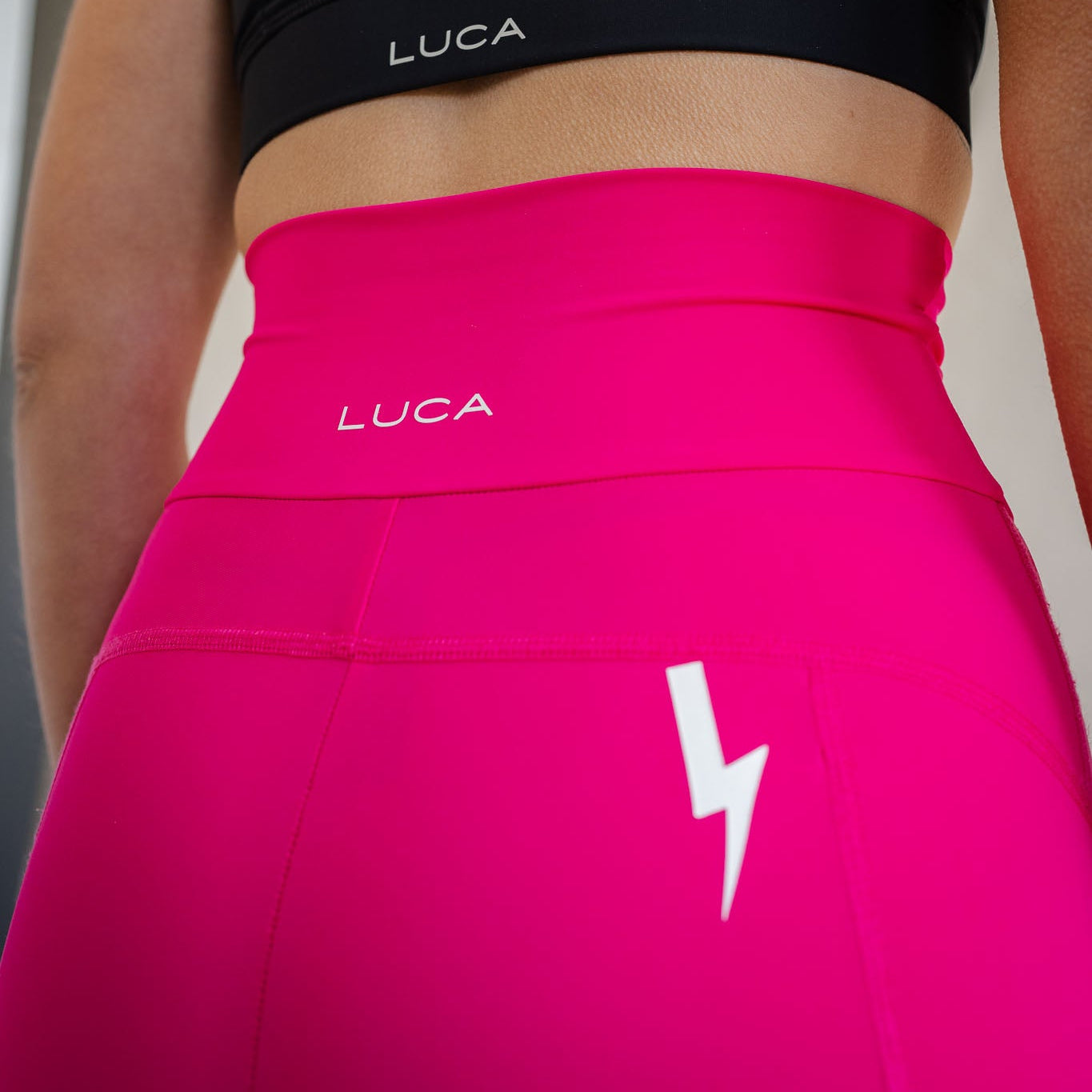 Close-up rear view showing the design details of LUCA women's high-waisted hot pink cycling shorts. Displays a white LUCA text logo centered on the wide waistband, flatlock stitching, and a signature white lightning bolt logo printed on the back right side.
