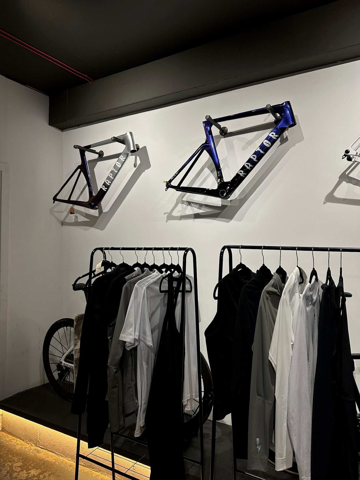 An-interior-photograph-of-a-retail- space-likely-a-cycling-urban-sports- brand-The-white-walls-feature-two- mounted-bicycle-frames-for-display- one-in-gray-and-one-in-a-metallic- blue-both-with-the-word-"RAPTOR"- visible-on-the-downtube-Below-the- frames-there-are-two-black-clothing- racks-displaying-a-selection-of- minimalist-apparel-in-shades-of-black- white-and-gray-including-t-shirts-tanks-and-collared-shirts-A-bicycle-wheel-is- partially-visible-in-the-bottom-left- corner