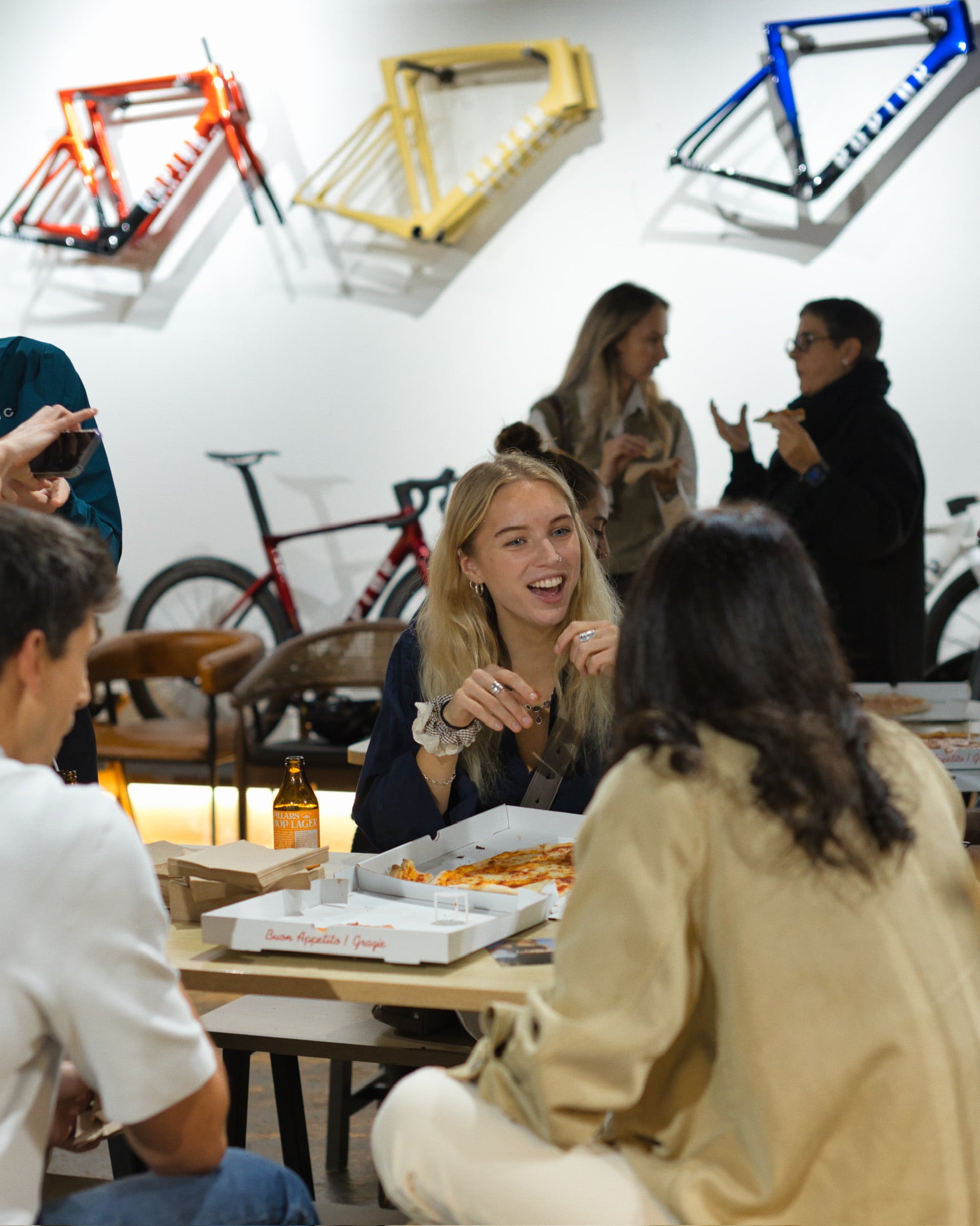 A-candid-indoor-photograph-of-a- diverse-group-of-young-adults- socializing-around-a-table-with-open- pizza-boxes-and-drinks-The-focus-is- on-a-blonde-woman-laughing-while- talking-to-a-friend-The-background- features-a-white-wall-with-three- brightly-colored-mounted-bicycle- frames-(red-yellow-and-blue)-and-a- complete-bicycle-visible-further-back- indicating-the-setting-is-a-cycling-shop-The-image-highlights- community-events-and-the-casual- atmosphere-around-the-cycling- brand