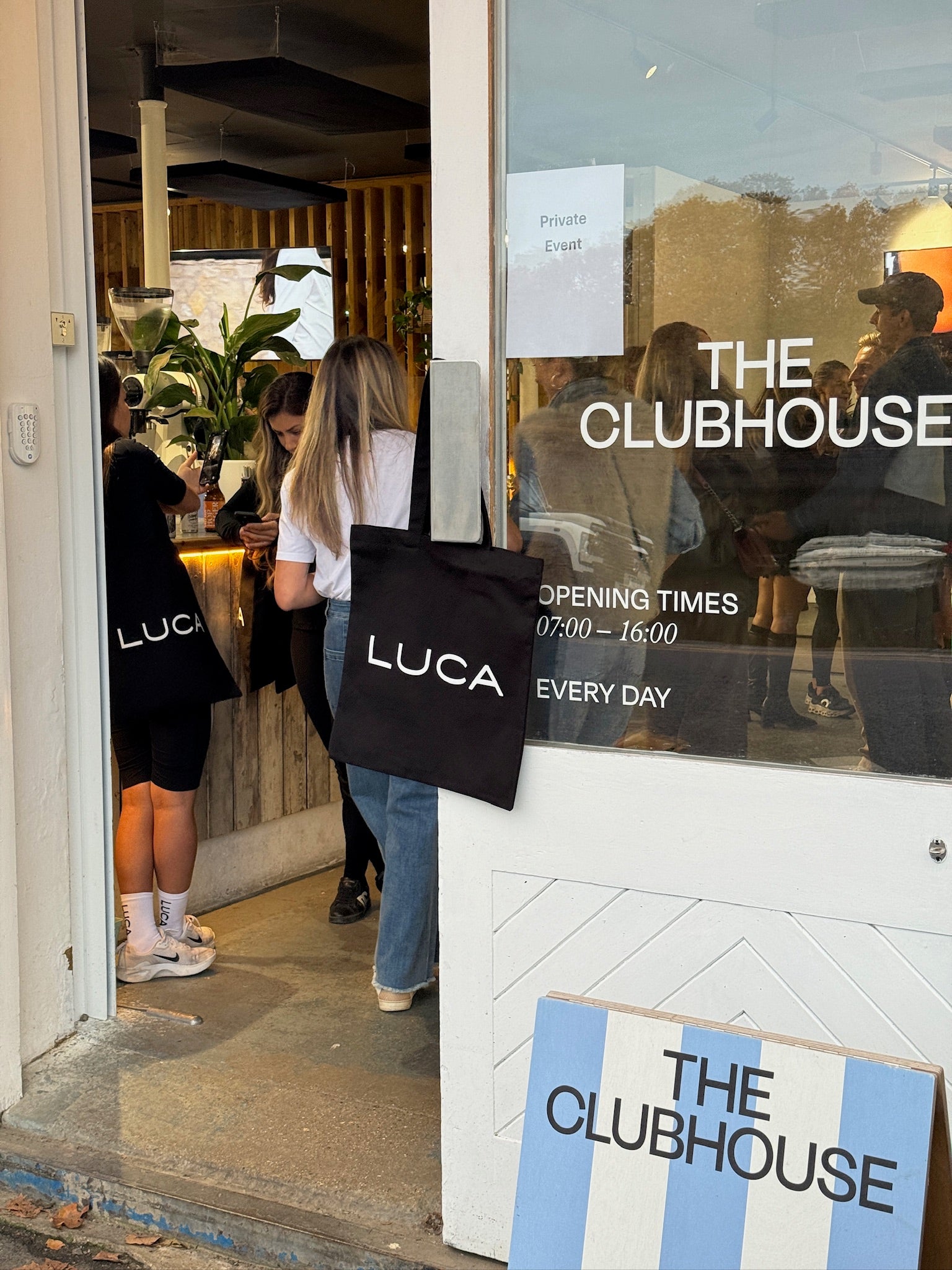 A-candid-photograph-of-people- entering-standing-in-the-doorway-of-a-storefront-labeled-"THE- CLUBHOUSE."-The-image-highlights- the-brand's-merchandise-a-woman's- back-is-visible-with-a-black-top-apron-featuring-the-white-text-"LUCA"-across-the-lower-back-and-another- person-is-carrying-a-black-canvas- tote-bag-with-the-white-"LUCA"- logo-The-image-conveys-brand- presence-community-event-and- physical-retail-experience-Store- opening-times-are-visible-on-the- glass-door