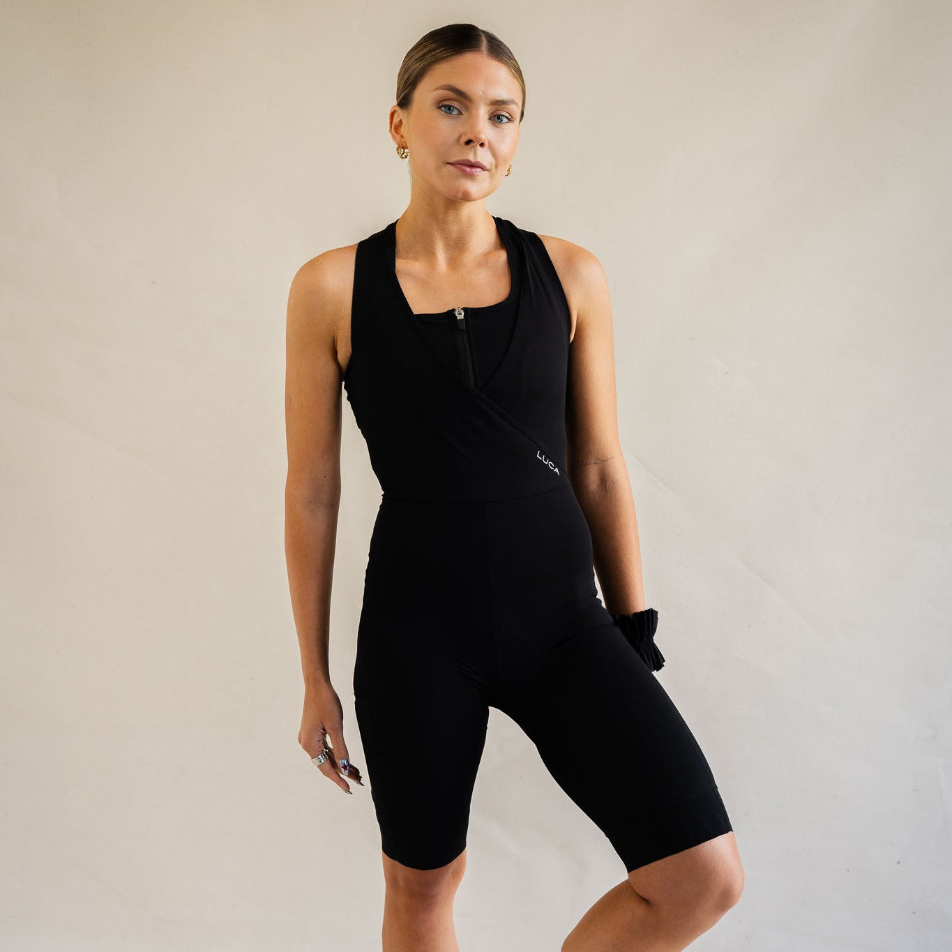 Front view of a woman modeling the LUCA women's premium cycling bib shorts in black. The image highlights the unique crossover V-neck bib strap design with a small white LUCA logo on the left rib, layered over a matching black front-zip sports bra, set against a plain studio background.