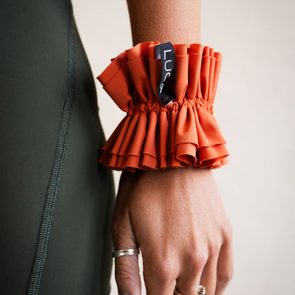 Close-up shot of a person wearing the LUCA Uplift Scrunchie in 'Aperol' orange on their wrist. The accessory features a unique, heavily ruffled tiered fabric design and a small black woven tag displaying the white LUCA logo, paired alongside dark olive green athletic wear.