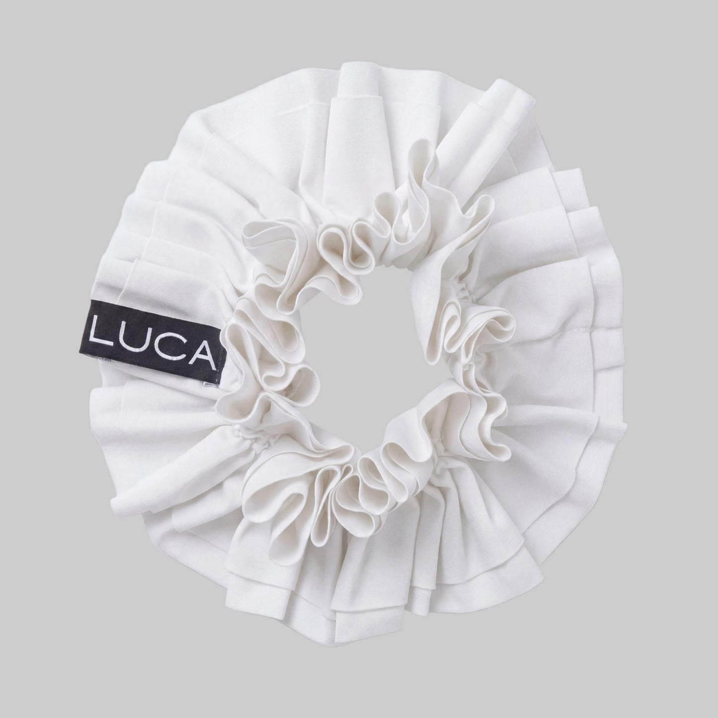 Flat-lay studio shot of the LUCA® Uplift Scrunchie in crisp Off White against a solid grey background. The accessory features a dramatic, multi-layered tiered ruffle fabric design and a prominent black woven tag displaying the white LUCA® brand name on the left side. - Off-White - 