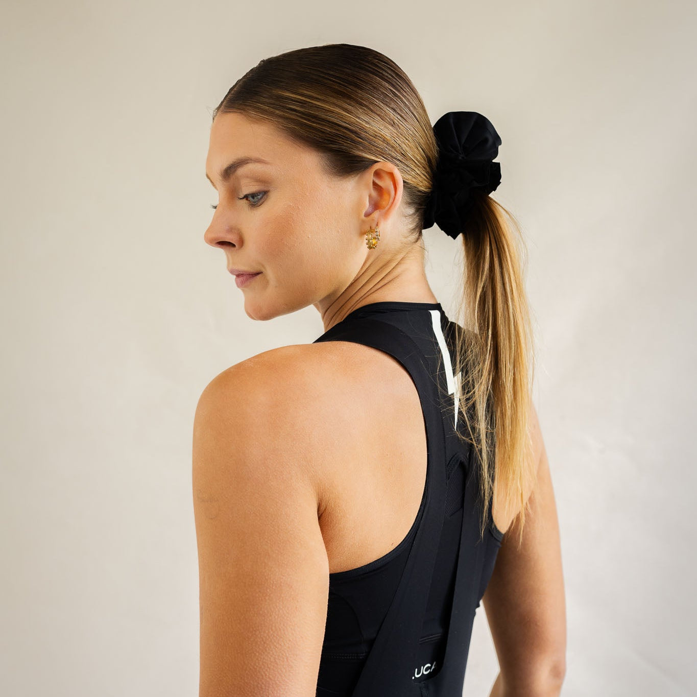 Side profile view of a woman with blonde hair in a ponytail secured by a black LUCA® Uplift ruffled hair scrunchie. She is wearing a black LUCA® racerback sports bra and black cycling bib shorts featuring a white lightning bolt graphic on the upper back strap, set against a neutral studio background.  