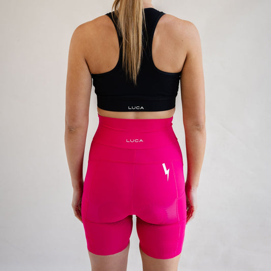 Rear view of a woman wearing a black LUCA racerback sports bra and high-waisted hot pink padded cycling shorts. Features white LUCA text logos centered on the bra band and waistband, with a white lightning bolt logo on the right thigh.