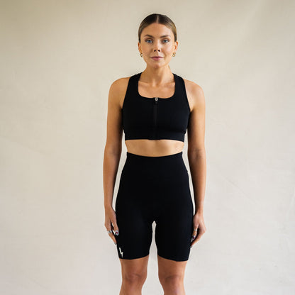 A front-facing studio shot of a woman looking forward against a cream background. She is wearing a matching black LUCA® athletic set, featuring a scoop neck sports bra top with a center front zipper and high-waisted cycling shorts. A small white lightning bolt graphic is visible on the lower right leg hem.