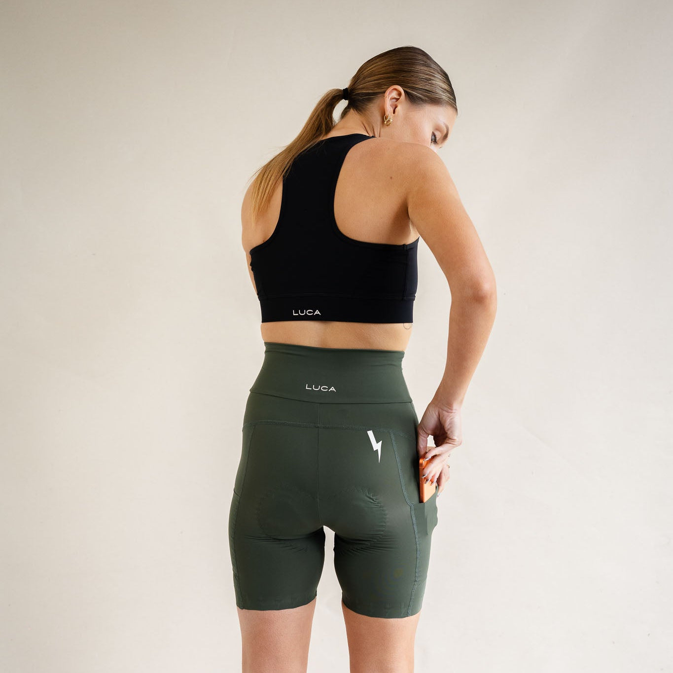 A rear view lifestyle shot of a woman looking over her shoulder against a cream background, sliding a smartphone into the side utility pocket of her high-waisted basil green LUCA® cycling shorts. She is wearing a matching black racerback sports bra, highlighting the white LUCA® logos on both pieces and a white lightning bolt graphic on her right hip. LUCA®