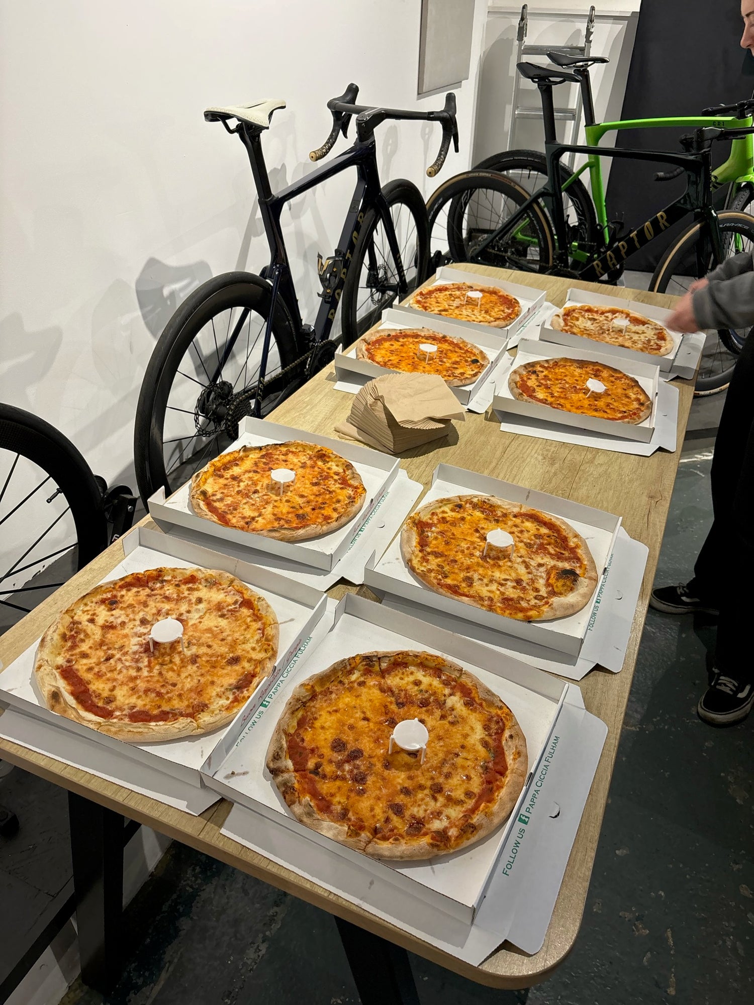 Several-whole-cheese-pizzas-laid- out-on-a-table-in-a-bike-cycling- event-with-high-end-bicycles-visible-in-the-background