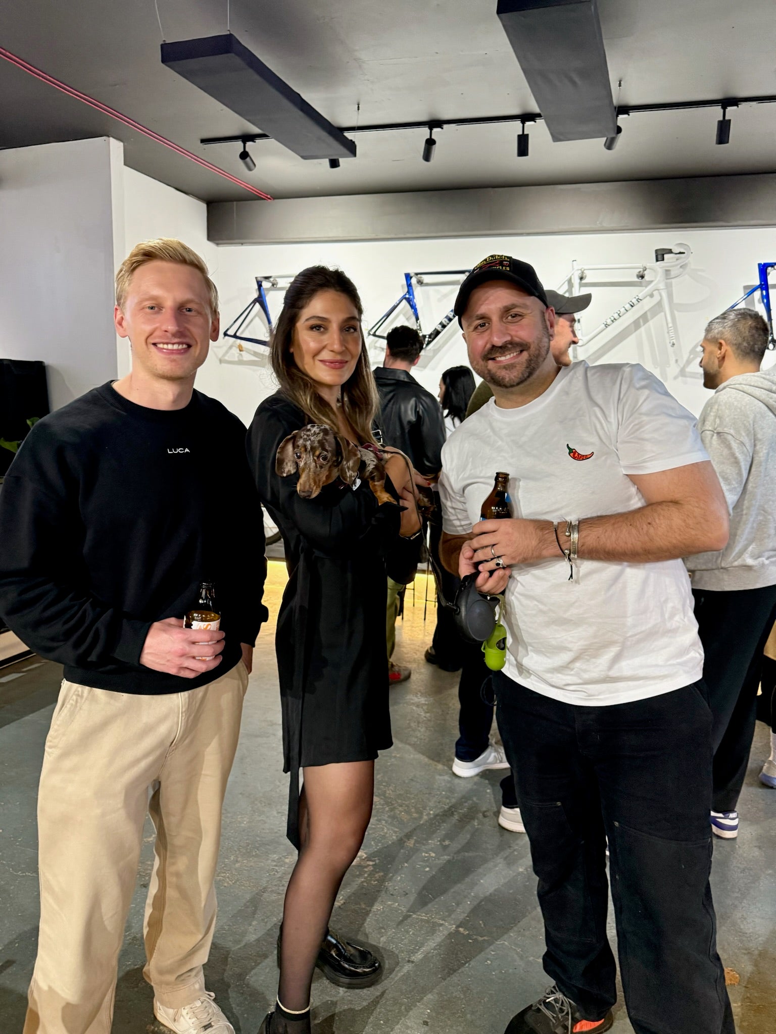 Three-friends-smiling-and-holding- drinks-at-an-indoor-event-with-bike- frames-decorating-the-wall-behind- them