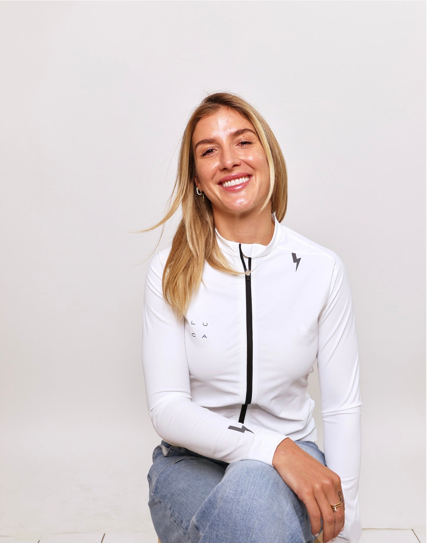 Studio-portrait-of-a-smiling-woman- with-blonde-hair-seated-and-wearing-a-white-long-sleeve- performance-top-cycling-jersey-that-features-a-black-full-length- zipper-and-the-LUCA-logo-printed-in-black-on-the-chest-A-small-black-lightning-bolt-logo-is-visible- on-the-shoulder