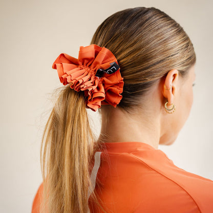 Rear view of a woman with blonde hair styled in a ponytail, secured by the LUCA Uplift Scrunchie in 'Aperol' orange. The shot highlights the voluminous, multi-layered ruffled fabric design and a small black tag with white branding, paired with a matching orange athletic top.