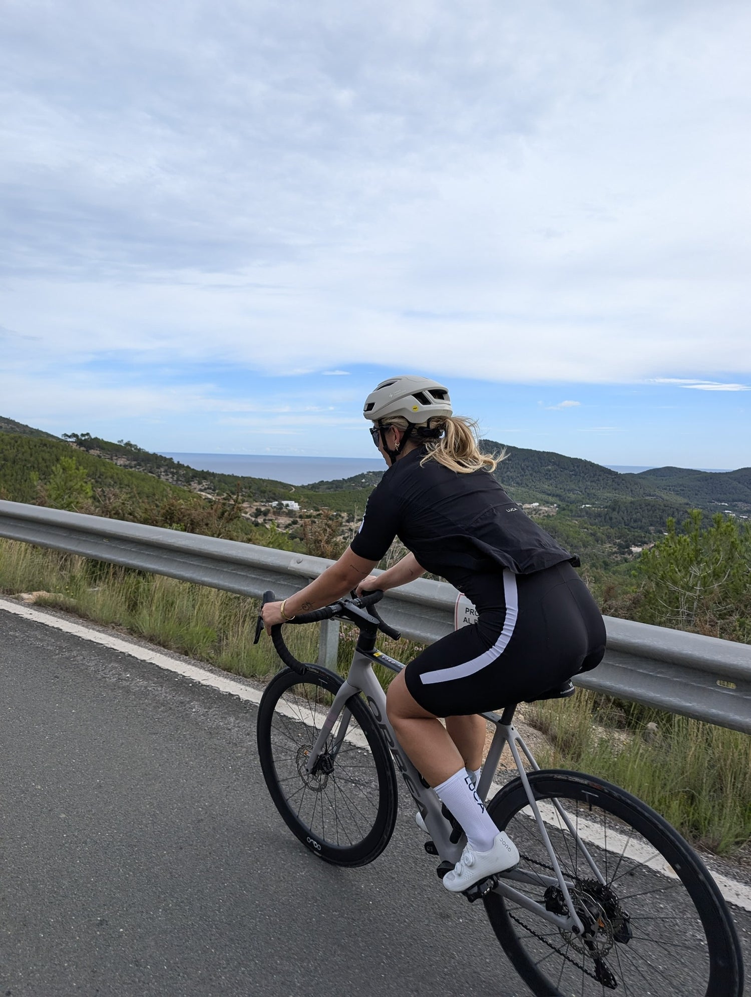A-female-cyclist-rides-a-grey-road-bike-along-a-coastal-road-with-a-metal-guardrail-overlooking-hills-and- the-ocean-under-a-partially-cloudy- sky-She-is-wearing-a-black-cycling-kit-with-white-stripes-on-the-bib- shorts-a-light-helmet-and- white-cycling-socks