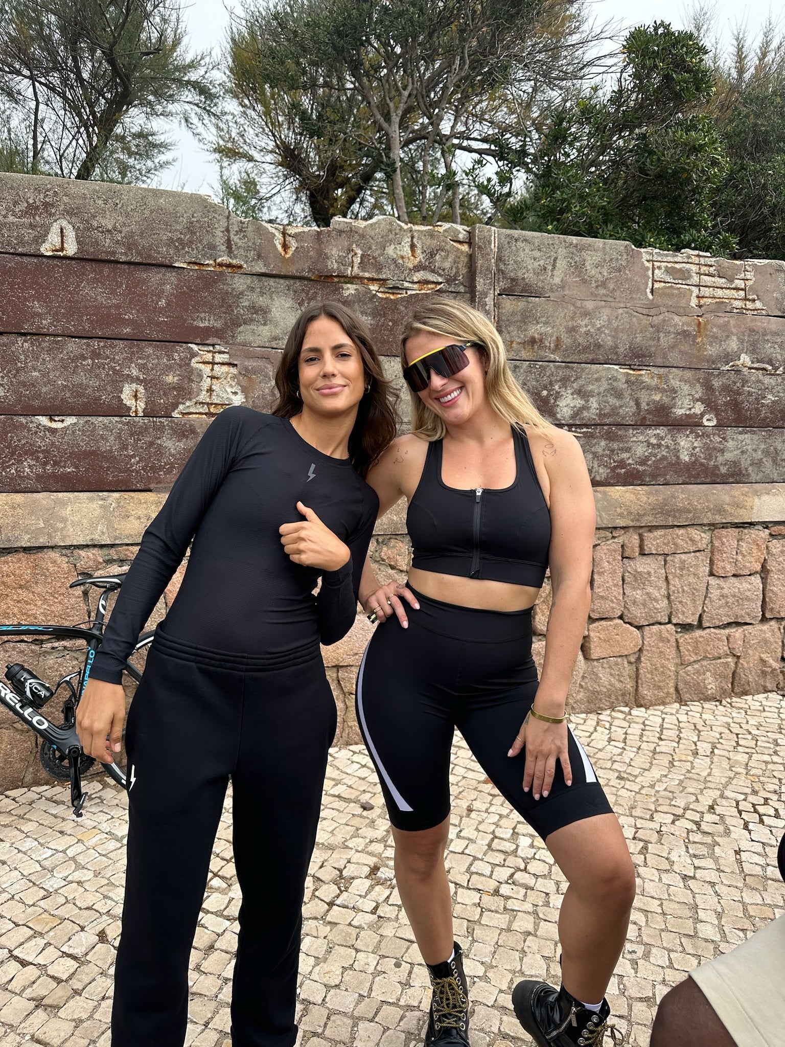 Two-women-posing-outside-in- matching-black-sportswear-one- wearing-a-long-sleeve-set-and-the- other-wearing-a-sport-bra-and-bike- shorts