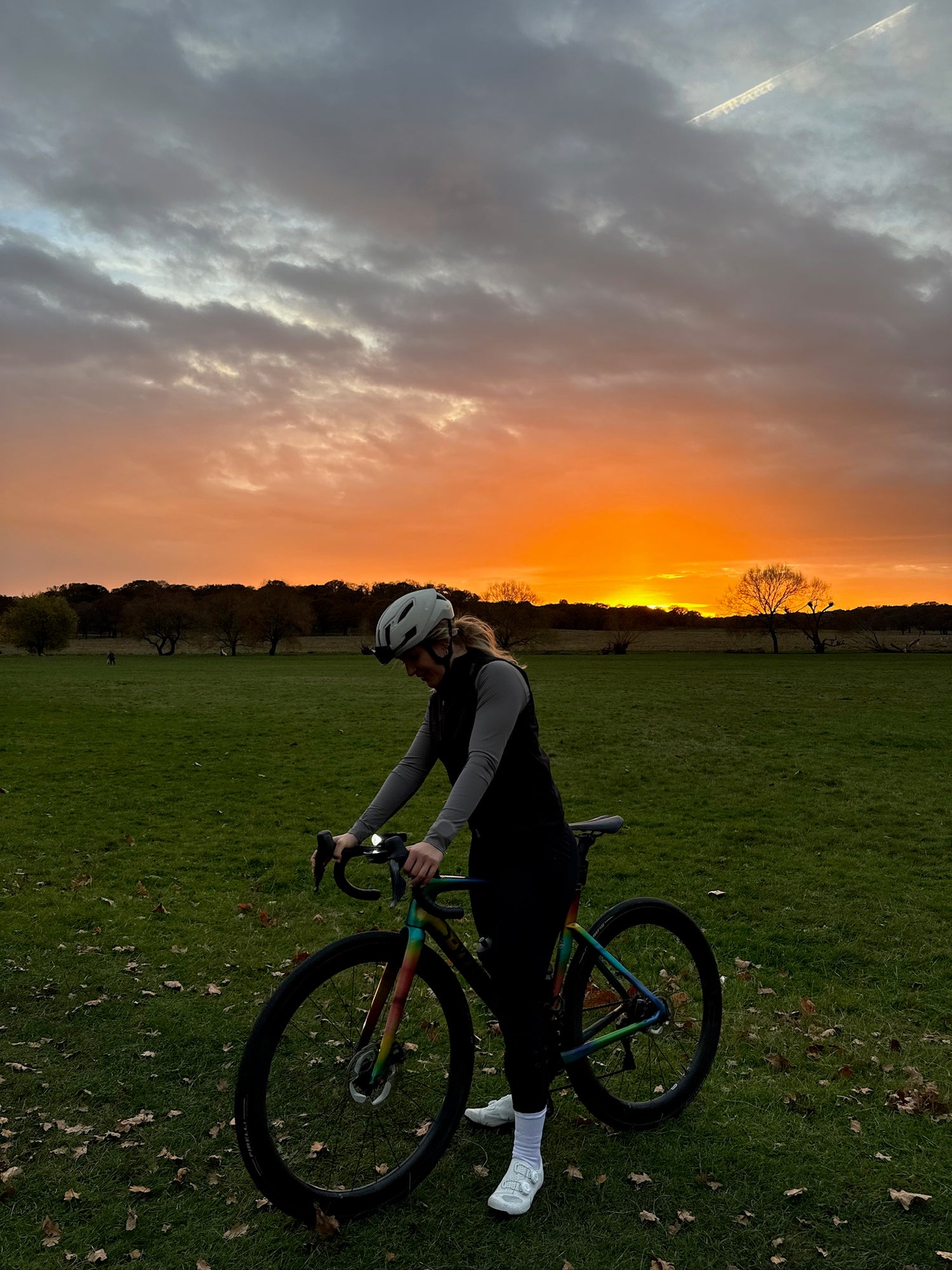 Woman-cyclist-standing-next-to-her- colorful-bike-in-a-park-at-sunset-, wearing-dark-apparel-and-a-helmet