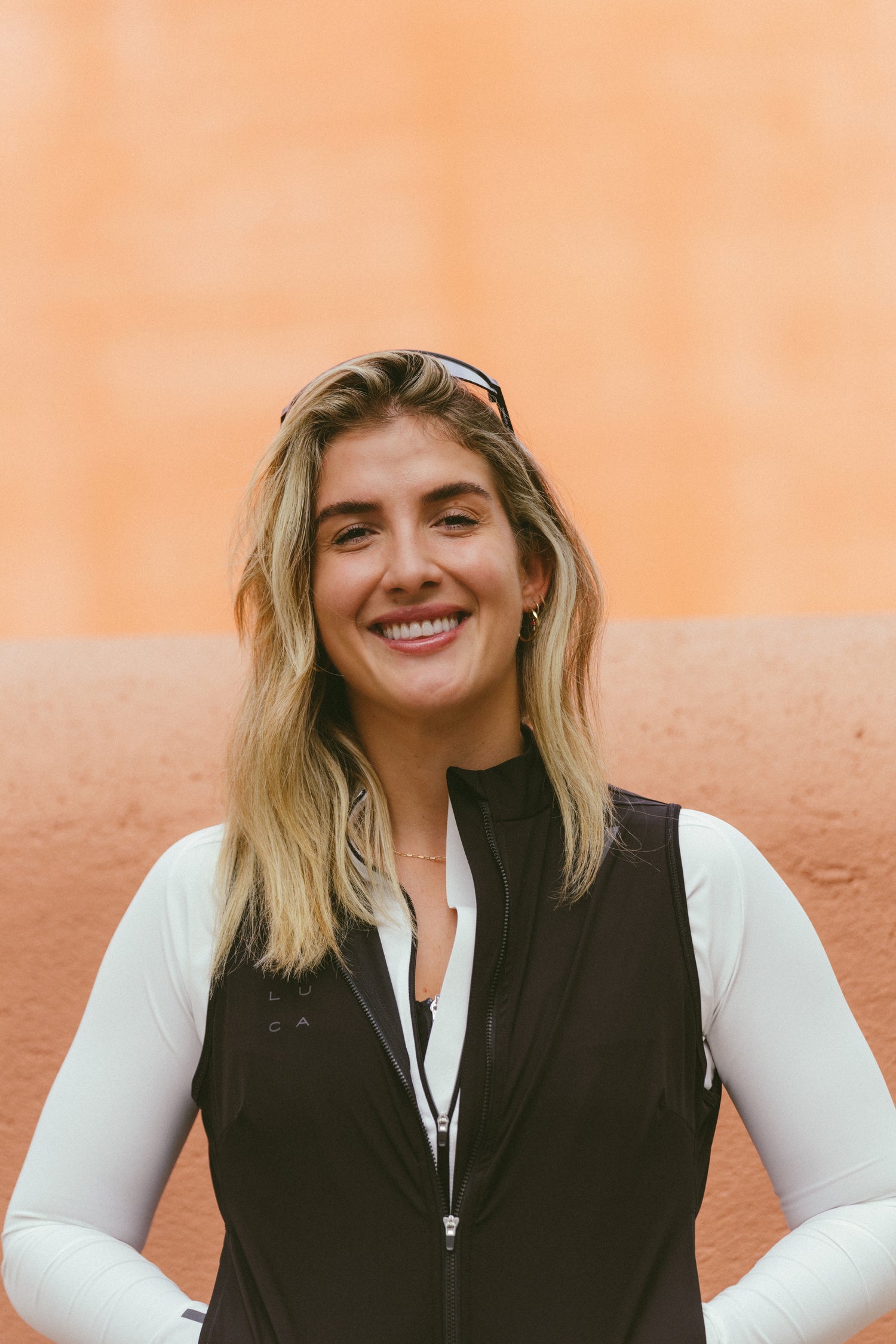 Smiling-woman-wearing-a-black-zip-up-vest-over-a-white-shirt-with- sunglasses-on-her-head-against-a- peach-background