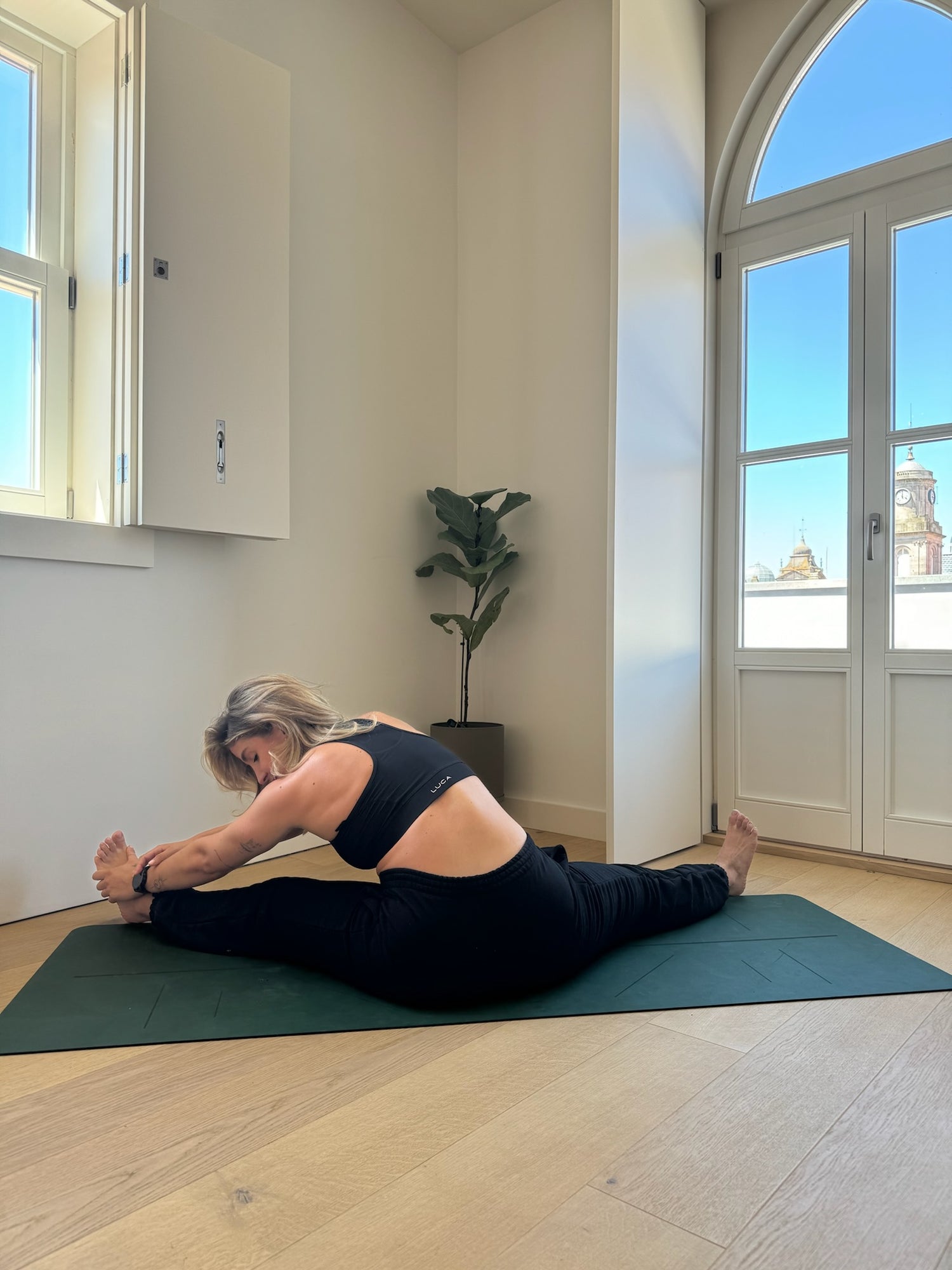 Woman-wearing-black-athletic- leggings-and-a-black-sports-bra- practicing-a-seated-straddle-stretch-Upavistha-Konasana-on-a- dark-green-yoga-mat