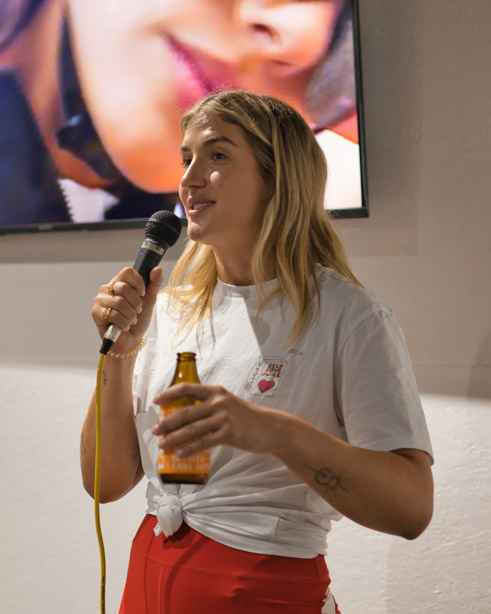 Woman-with-blonde-hair-wearing-a- white-t-shirt-and-red-pants-giving-a-presentation-talk-holding-a- microphone-and-a-glass-bottle