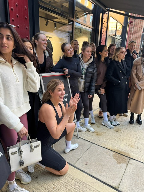 A-large-group-of-women-gathered- excitedly-outside-a-storefront-likely- for-an-event-product-launch-The- focus-is-on-a-woman-in-the- foreground-who-is-kneeling-and- clapping-while-wearing-black- activewear-and-white-socks-Many- attendees-are-wearing-casual- athletic-winter-street-style-apparel