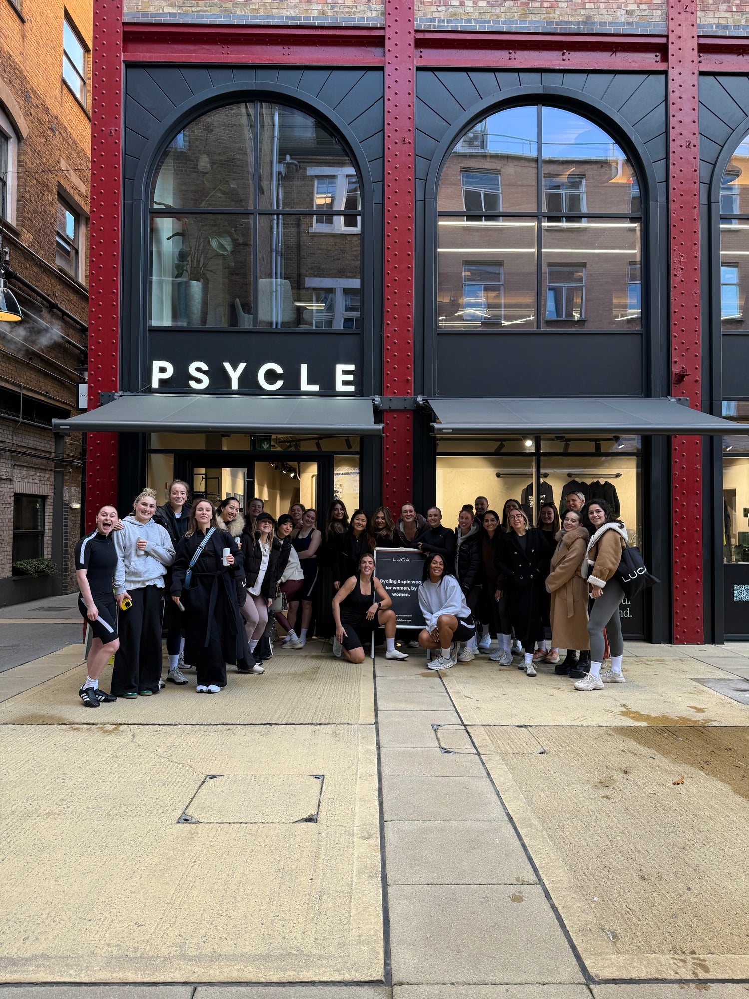 A-large-group-of-people-mostly- women-gathered-enthusiastically- outside-the-entrance-of-a-fitness- studio-named-"PSYLE,"-which-features-a-distinctive-red-brick-and- black-steel-facade-with-arched- windows-Many-in-the-group-are- wearing-black-casual