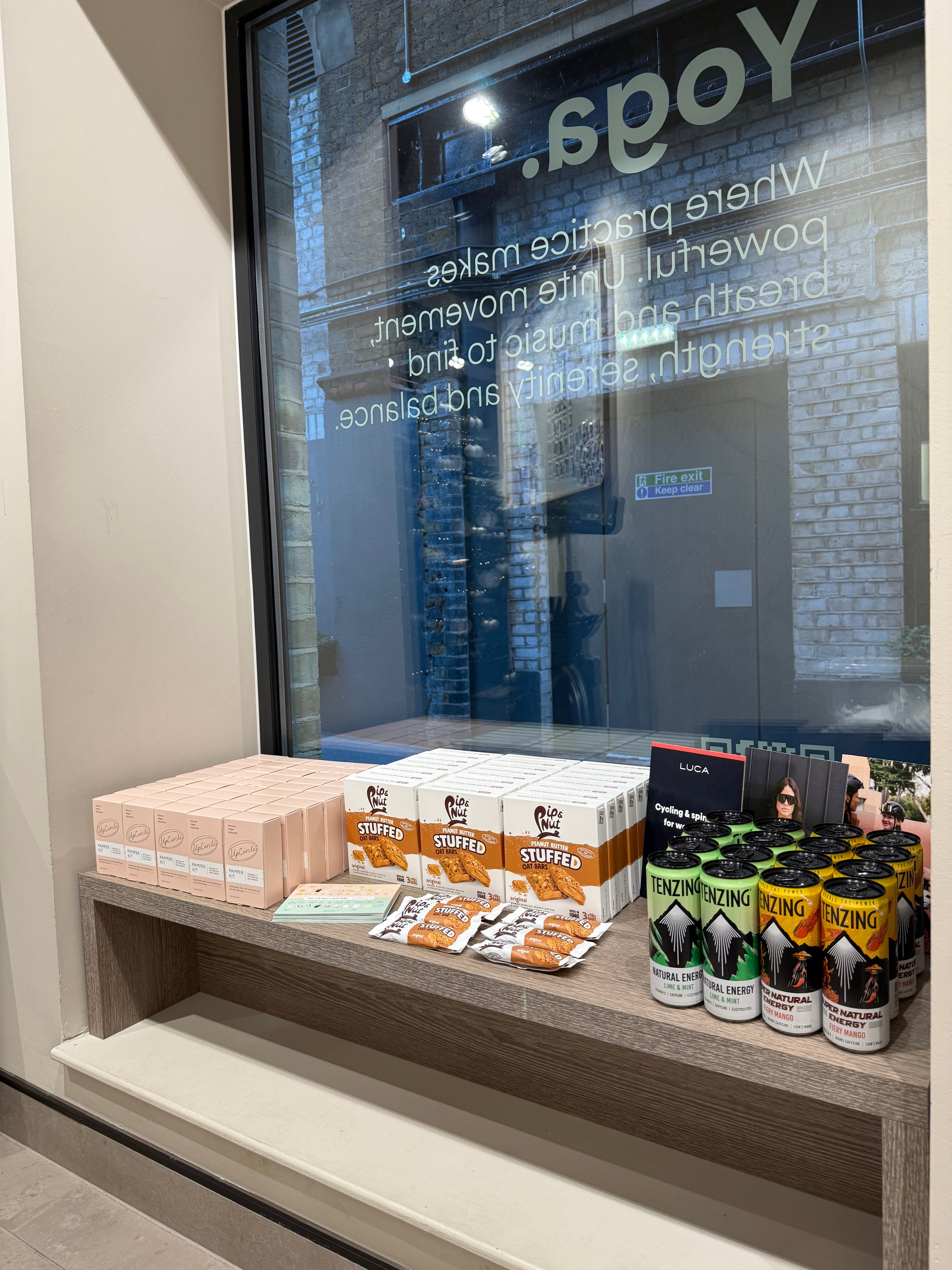 Retail-display-of-pre-and-post-workout-snacks-and-drinks-set-up- in-a-studio-window-including-stacks-of-pink-boxes-white-boxes-of-Stuffer-orange-snack-packs-and- cans-of-Tenzing-energy-drinks-Text- reflecting-in-the-window-advertises- Yoga
