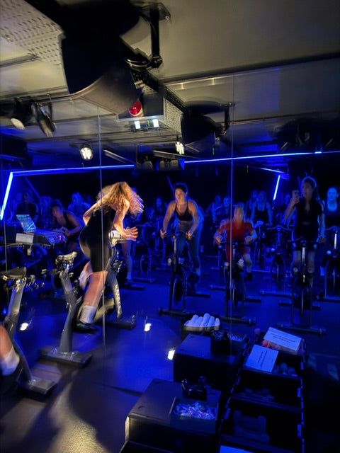 Indoor-cycling-studio-during-a-class-featuring-approximately-a- dozen-people-on-stationary-bikes-in-a-dark-room-illuminated-by- vibrant-blue-neon-strip-lights-The- focus-is-on-a-woman-in-the- foreground-with-long-blonde-hair- standing-up-on-her-bike-and- vigorously-cycling-The-atmosphere- is-energetic-and-dynamic
